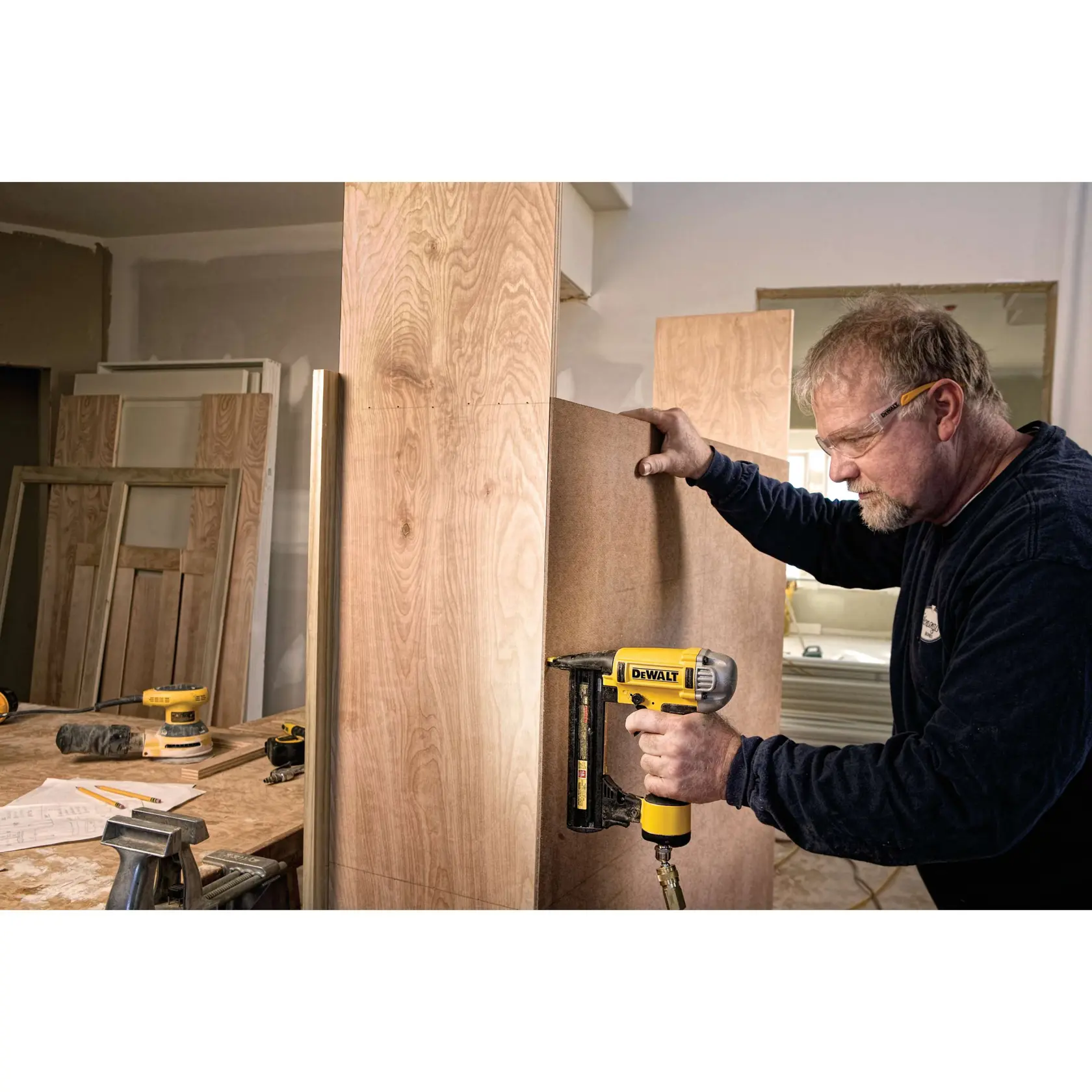 A person using a DEWALT DWFP1838 pneumatic nailer to secure a wood panel during a carpentry project. The individual's face is blurred, and there are various woodworking tools and materials visible in the workshop.