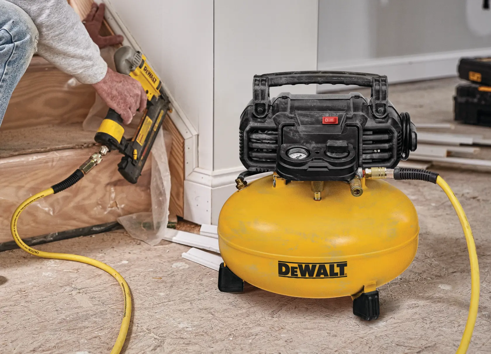 A DEWALT air compressor is connected to a pneumatic nail gun, being used to install trim along a staircase. The compressor and nail gun are yellow and black, and are part of a woodworking project.