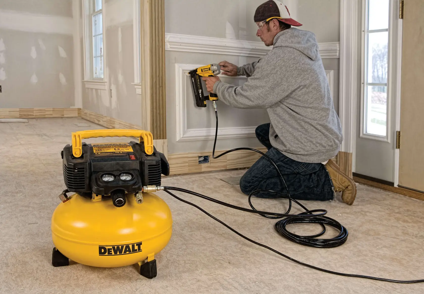 A person kneeling on the floor is using a DEWALT air compressor connected to a pneumatic nail gun to install trim on an interior wall in a room under construction.