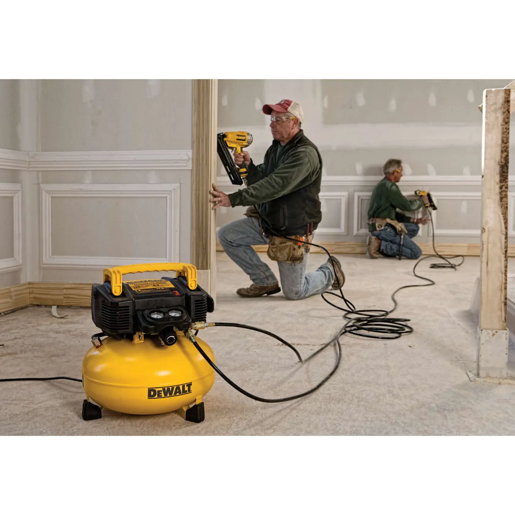 A DEWALT DWFP55126 yellow pancake air compressor is shown in the foreground of a construction site. Two workers are installing trim and molding using pneumatic nail guns connected to the compressor. The workspace has unfinished walls and baseboards.