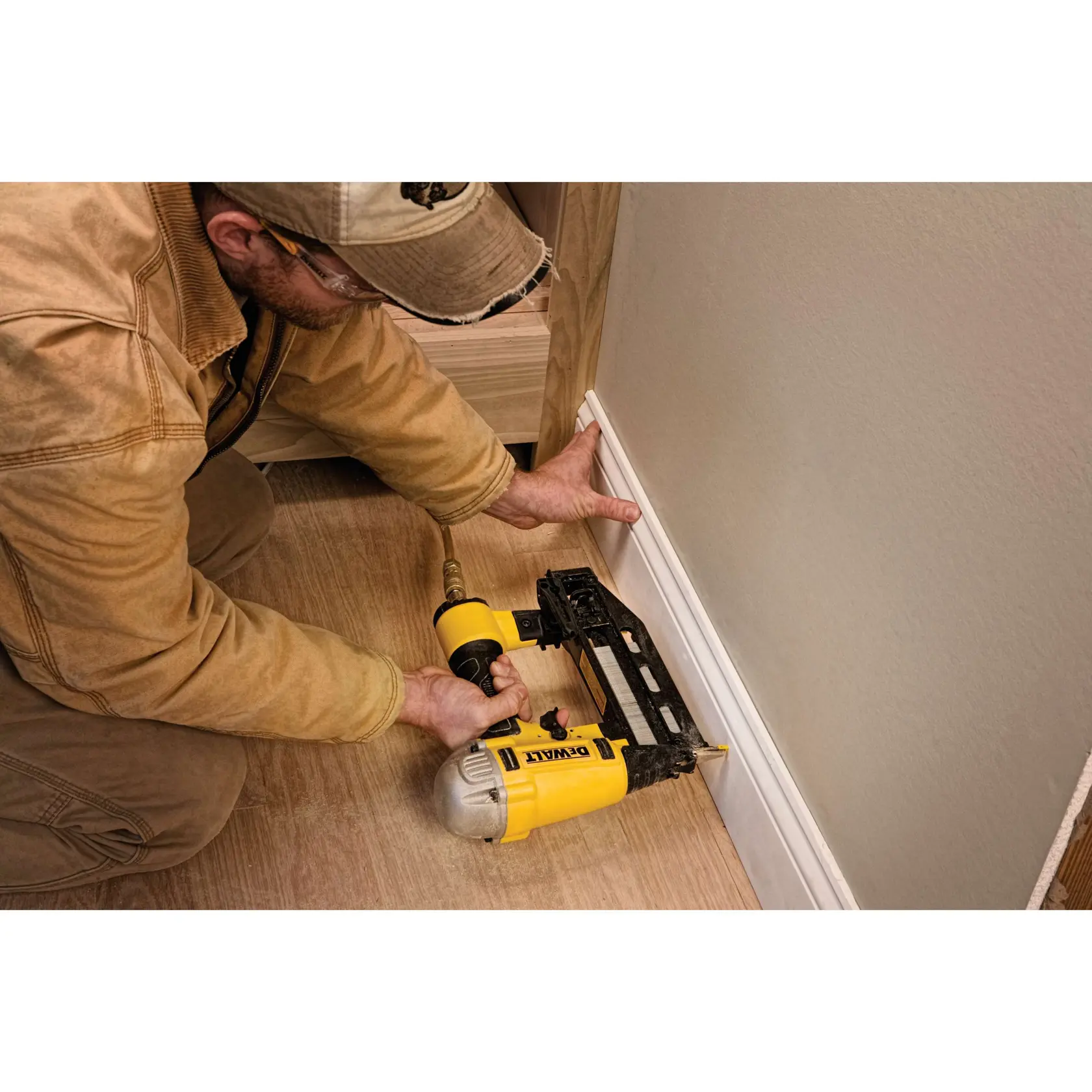 A person is using a yellow DEWALT pneumatic nailer to secure a baseboard to the wall. The person is kneeling on the floor and holding the nailer against the white trim.
