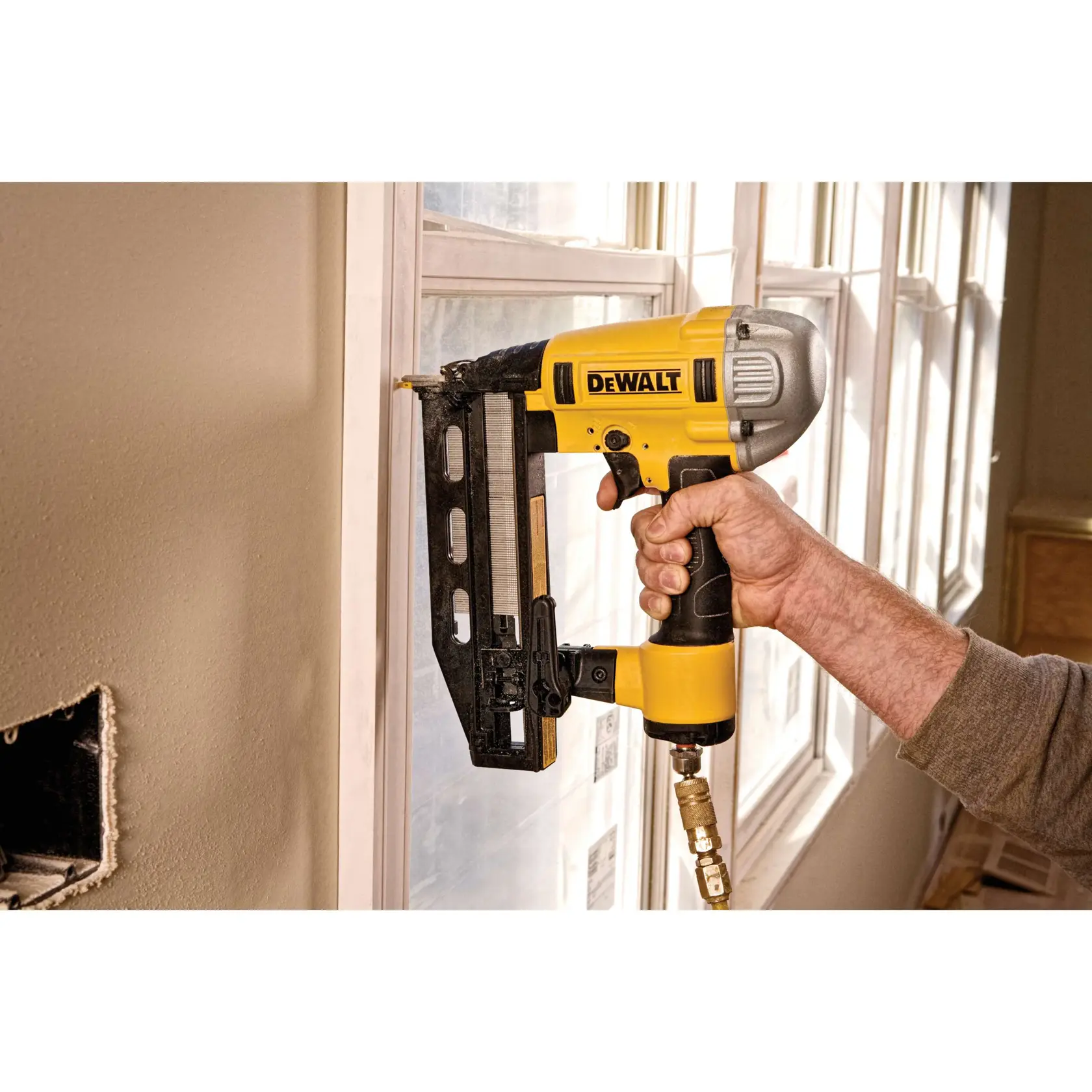A person using a yellow and black DEWALT pneumatic nail gun to attach trim to a window frame inside a room.