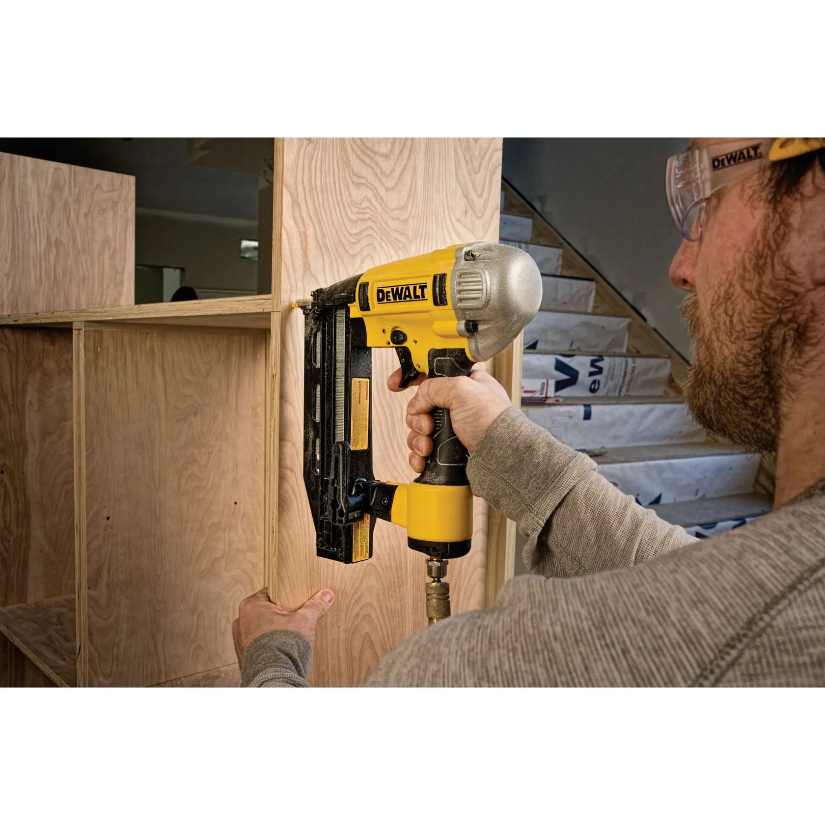 A DEWALT pneumatic nailer being used to assemble wooden cabinetry in a construction setting. The tool is yellow and black, and is connected to an air hose. The user’s hands are visible, holding the nailer against the wood.