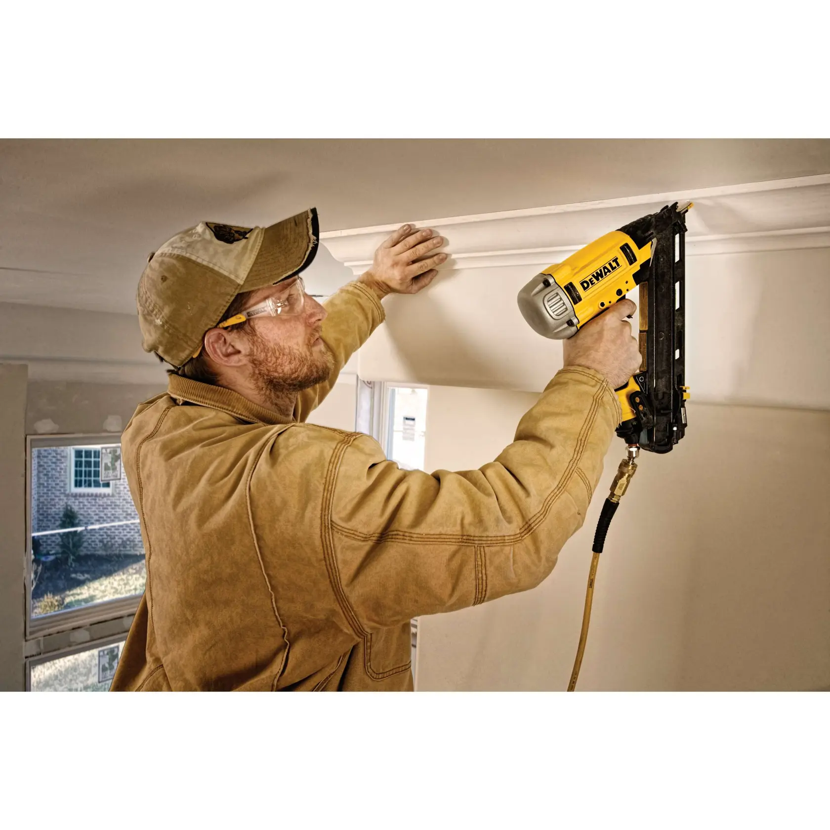 A person using a DEWALT DWFP72155 pneumatic nailer to install crown molding on the ceiling. The person is wearing a tan jacket and a tan cap, and the workspace is inside a room with windows visible.