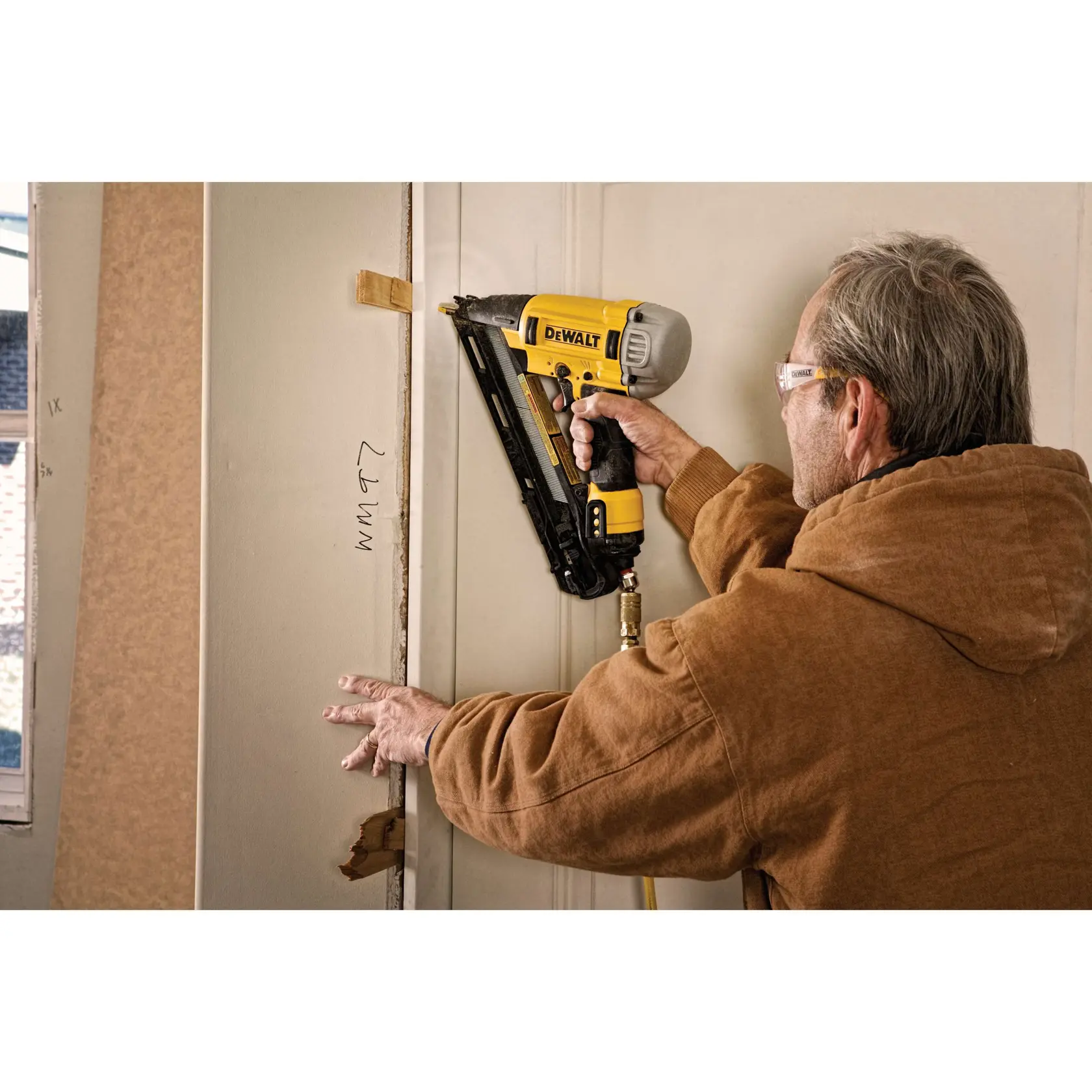 A person using a DEWALT pneumatic nail gun to attach trim or molding to a doorframe. The person is wearing safety glasses and a brown jacket.