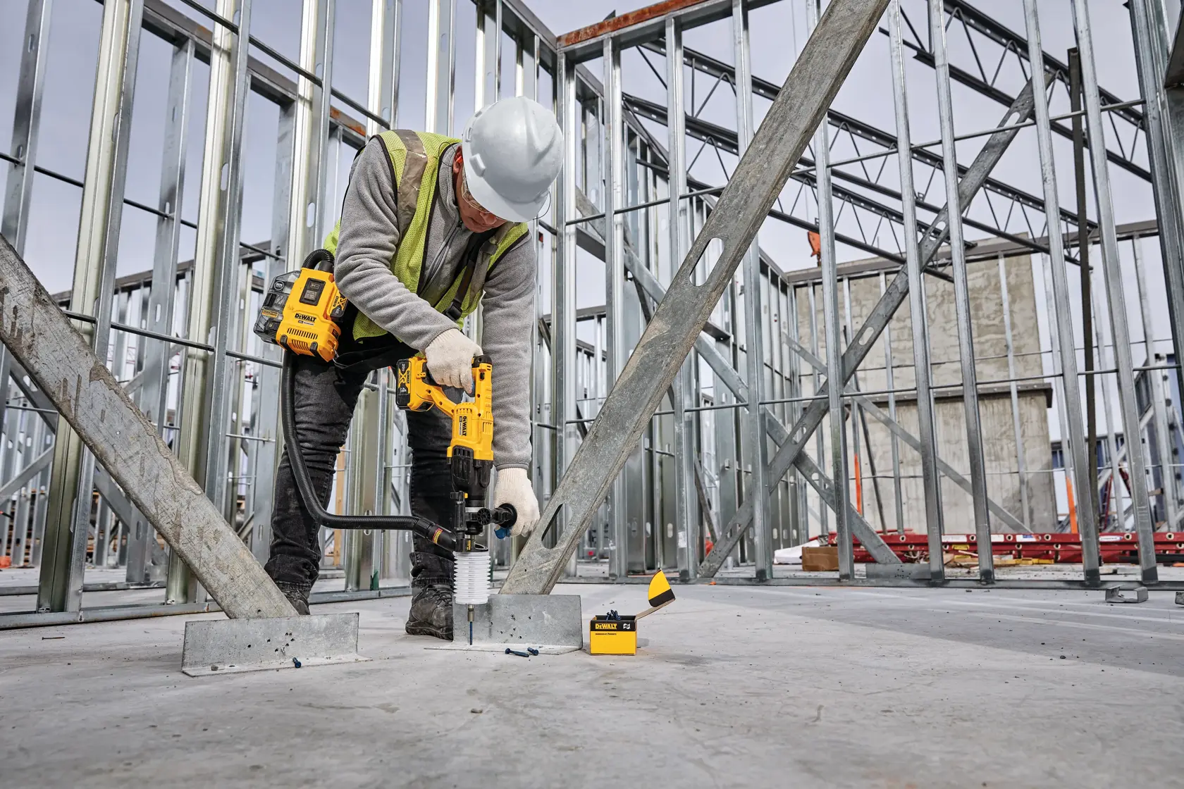 A construction worker wearing a hard hat and safety vest uses a yellow DEWALT power tool to drill into a concrete floor on a building site with metal framing.