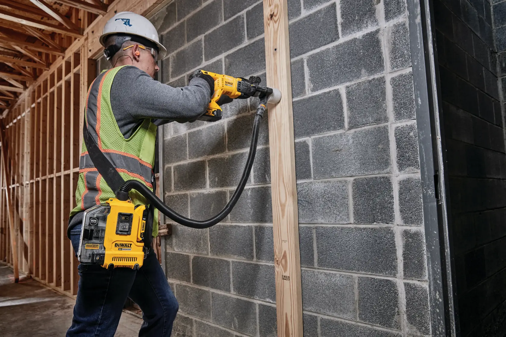 A construction worker uses a DEWALT cordless dust extractor with a power tool to drill into a wooden beam mounted on a cinder block wall. The worker is wearing a safety helmet, ear protection, and a safety vest.