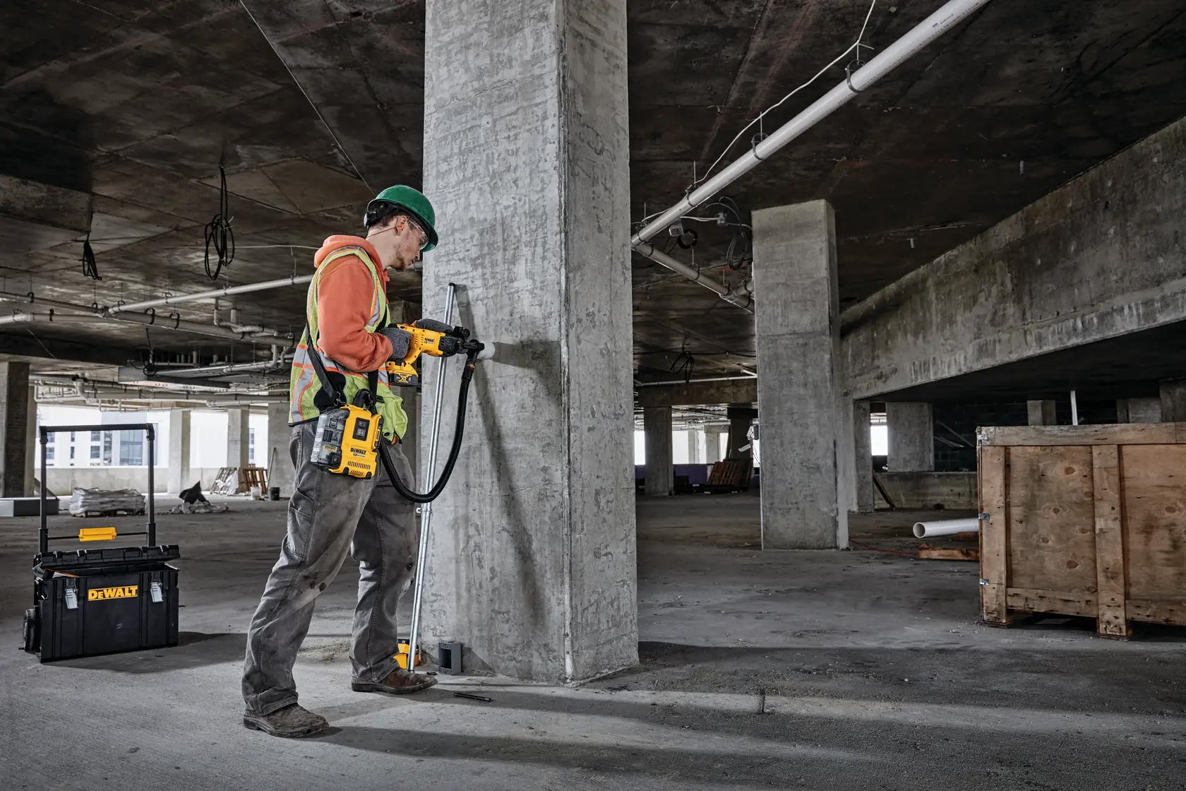 A construction worker wearing safety gear uses a DEWALT drill with an attached dust extractor to drill into a concrete pillar at a construction site. A DEWALT tool case is visible in the background.