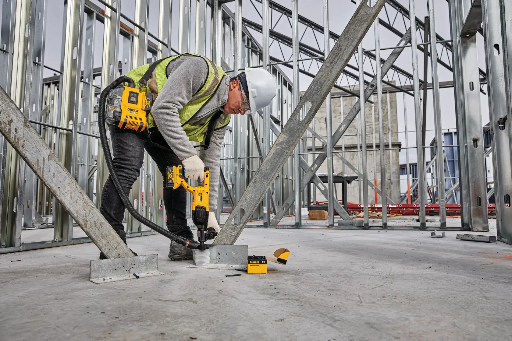 A construction worker wearing a safety helmet, gloves, and a reflective vest uses a DEWALT power tool to fasten metal supports to the concrete floor inside a building under construction. Metal framing and other construction materials are visible in the background.