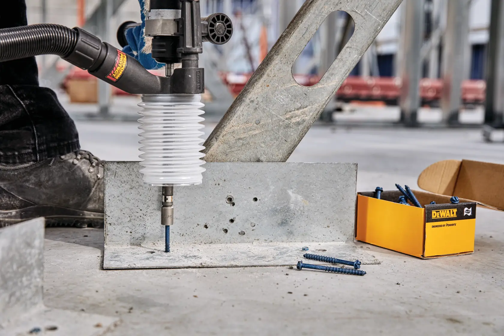 Close-up of a DEWALT dust extraction attachment being used with a power tool to drill into a metal plate on a construction site. A box of screws and loose screws are visible on the ground nearby.
