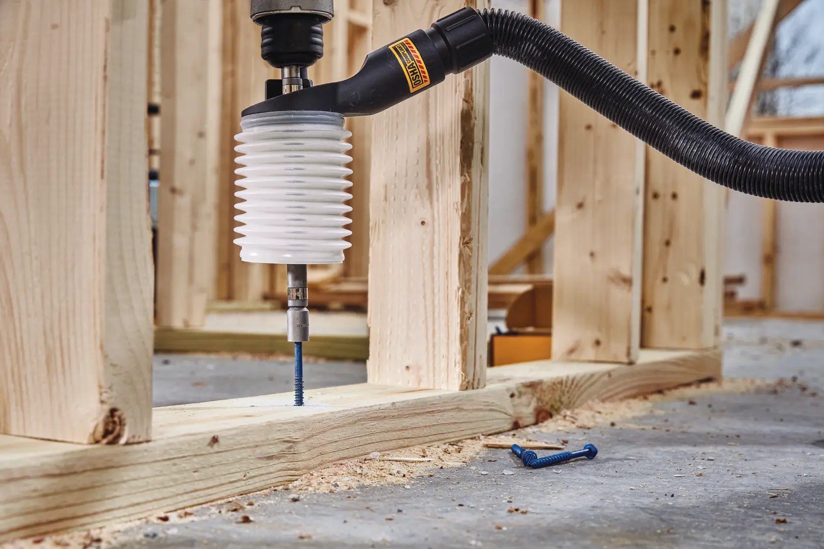 A DEWALT dust extraction attachment connected to a drill, being used to drill into wooden studs in a construction site. The attachment has an accordion-style white sleeve and is connected to a vacuum hose for dust collection.