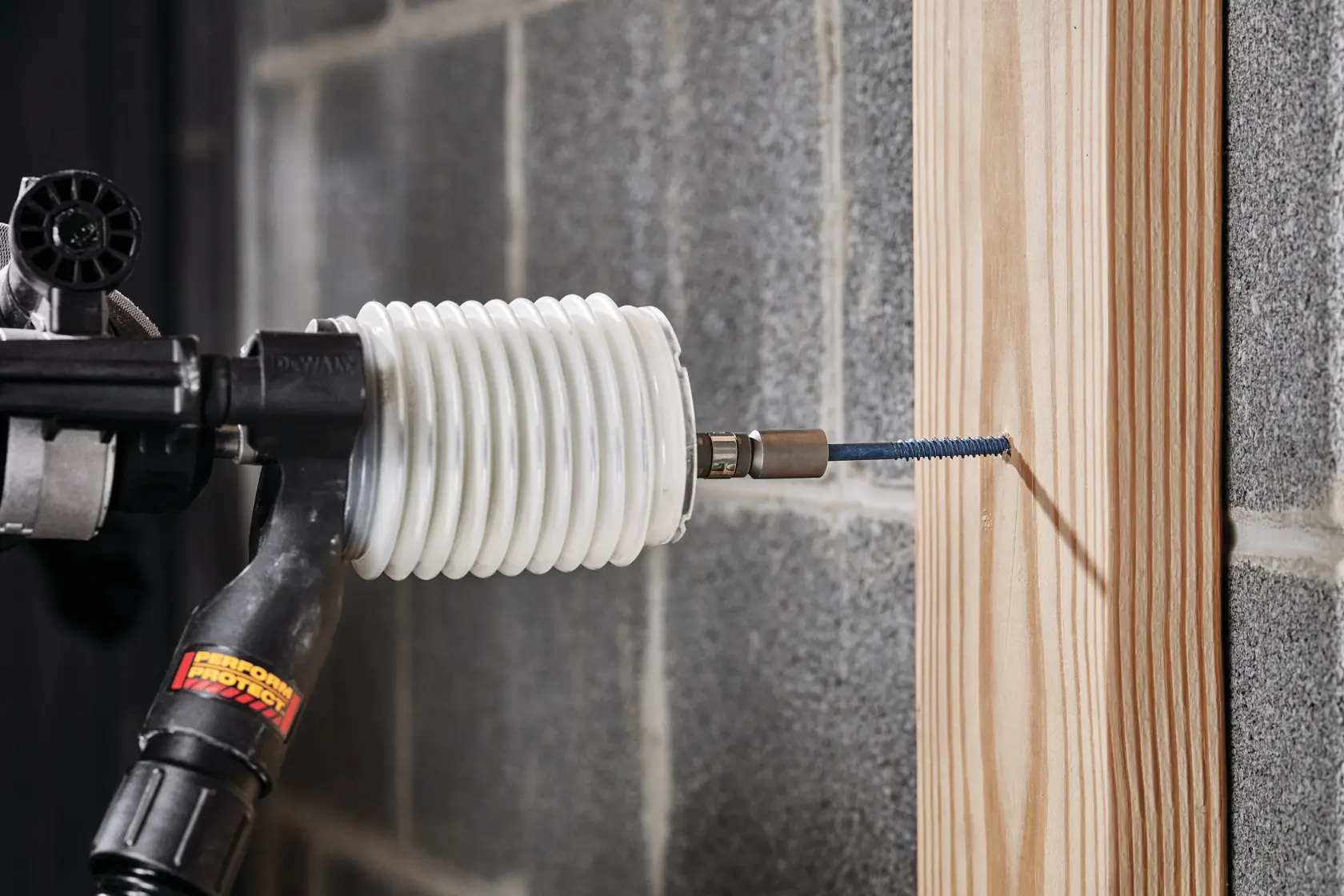 A DEWALT drill with a white dust extraction attachment is being used to drive a screw into a wooden beam mounted on a concrete block wall.