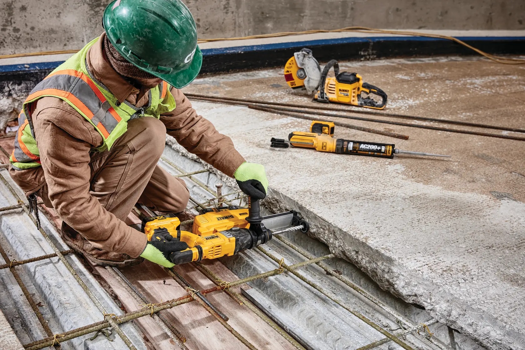 A construction worker wearing safety gear and a green hard hat is using a yellow DEWALT power tool to drill into a concrete slab. Additional DEWALT tools are visible on the floor, including a saw and a rotary hammer, all equipped for heavy-duty construction work.