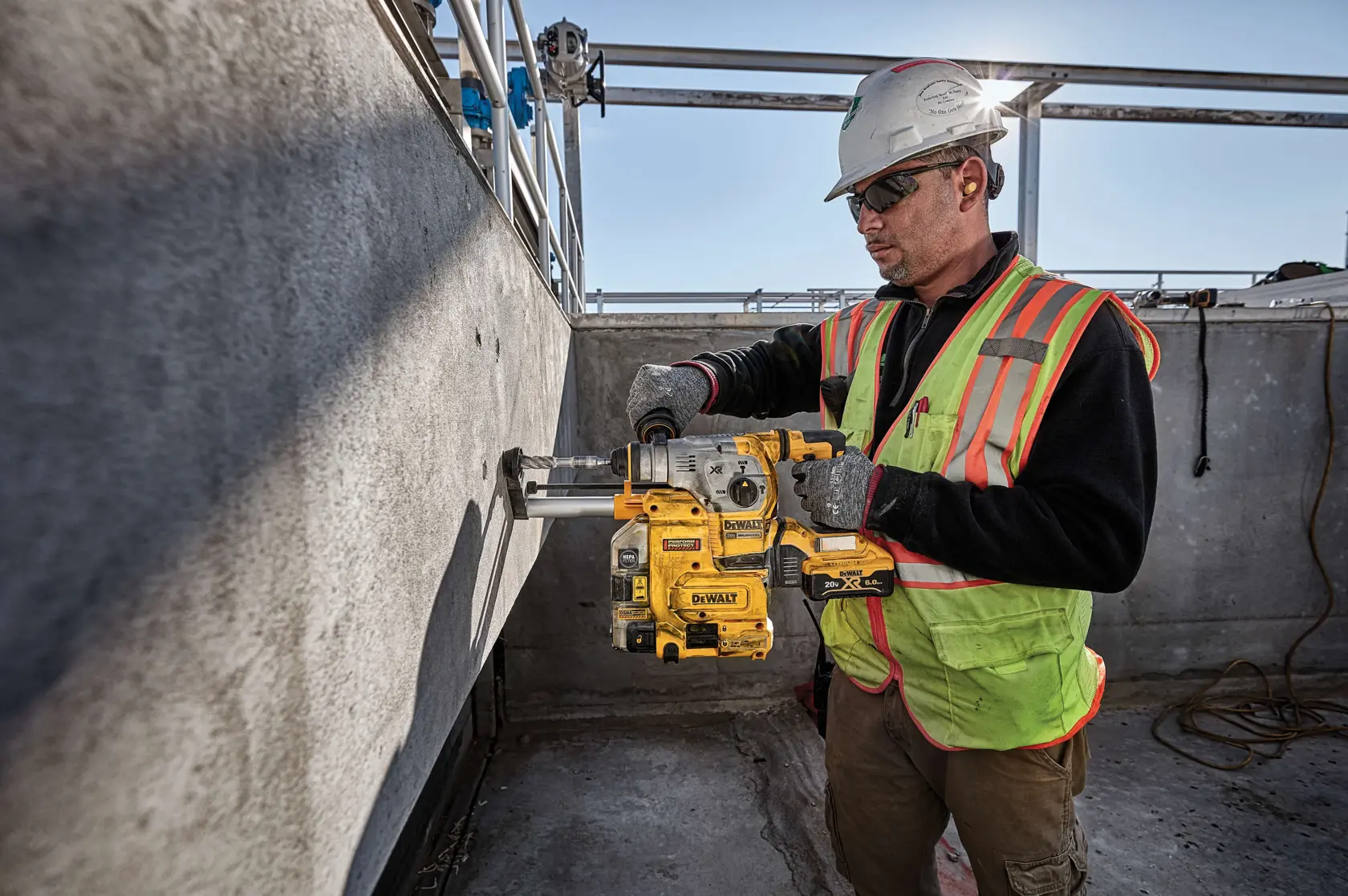 A construction worker wearing a safety helmet, gloves, and a high-visibility vest is using a yellow DEWALT DWH304DH tool to drill into a concrete wall at an outdoor construction site.