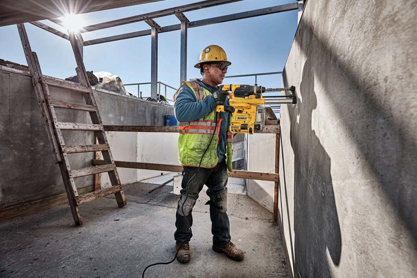 A construction worker wearing a yellow hard hat and safety vest uses a DEWALT power tool to drill into a concrete wall at a construction site with steel beams and a wooden ladder in the background.