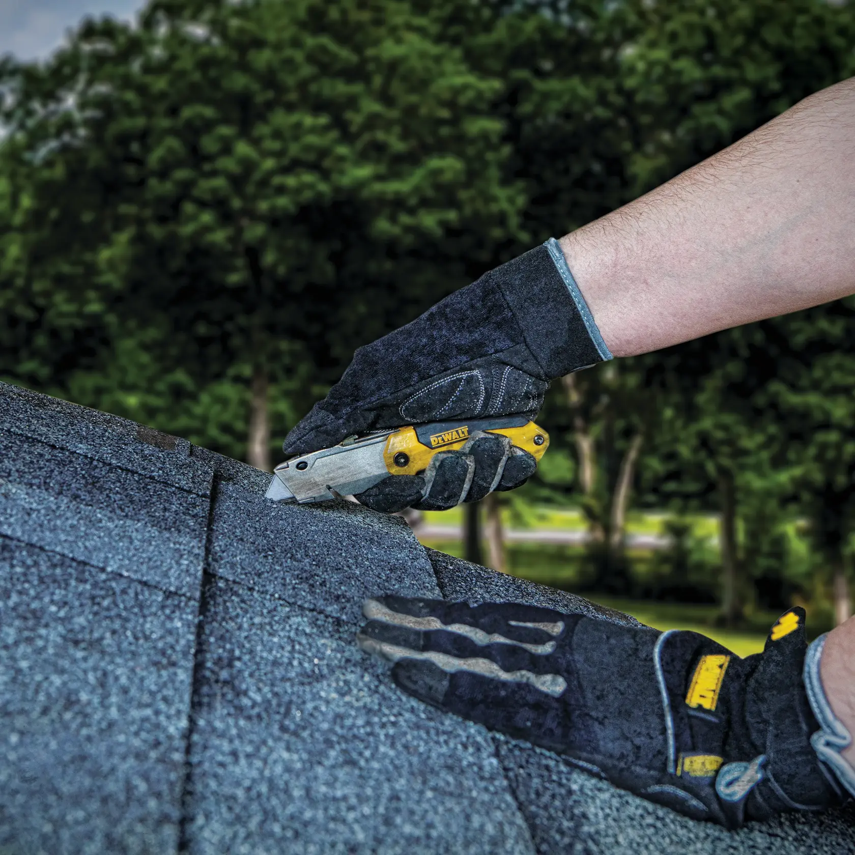 A person wearing gloves uses a DEWALT utility knife to cut roofing shingles outdoors.