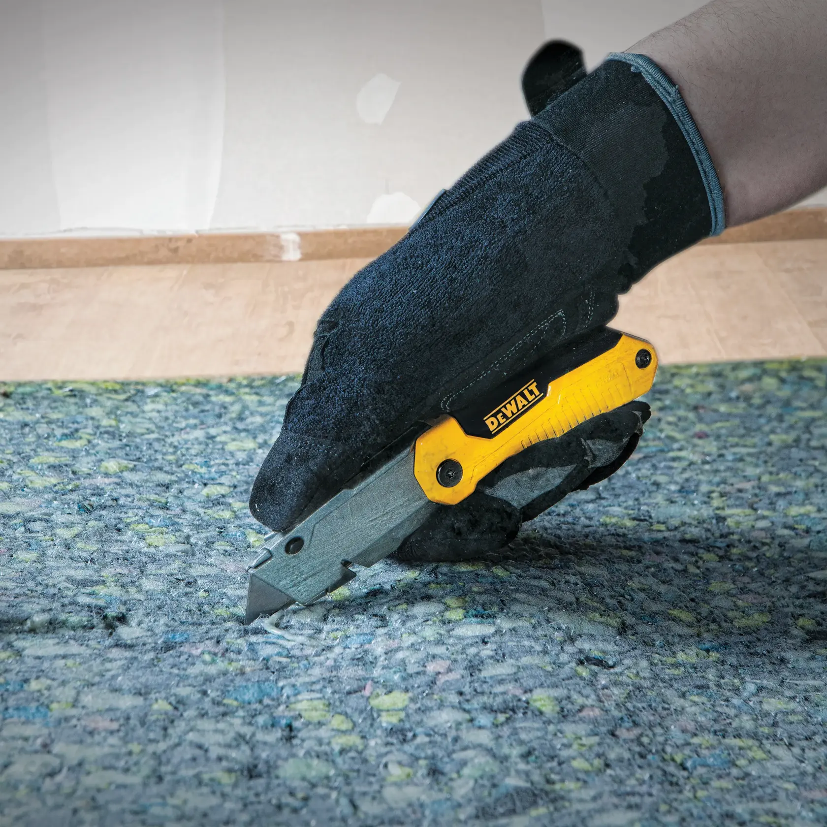 A person wearing a black glove uses a DEWALT utility knife to cut through carpet padding on the floor.