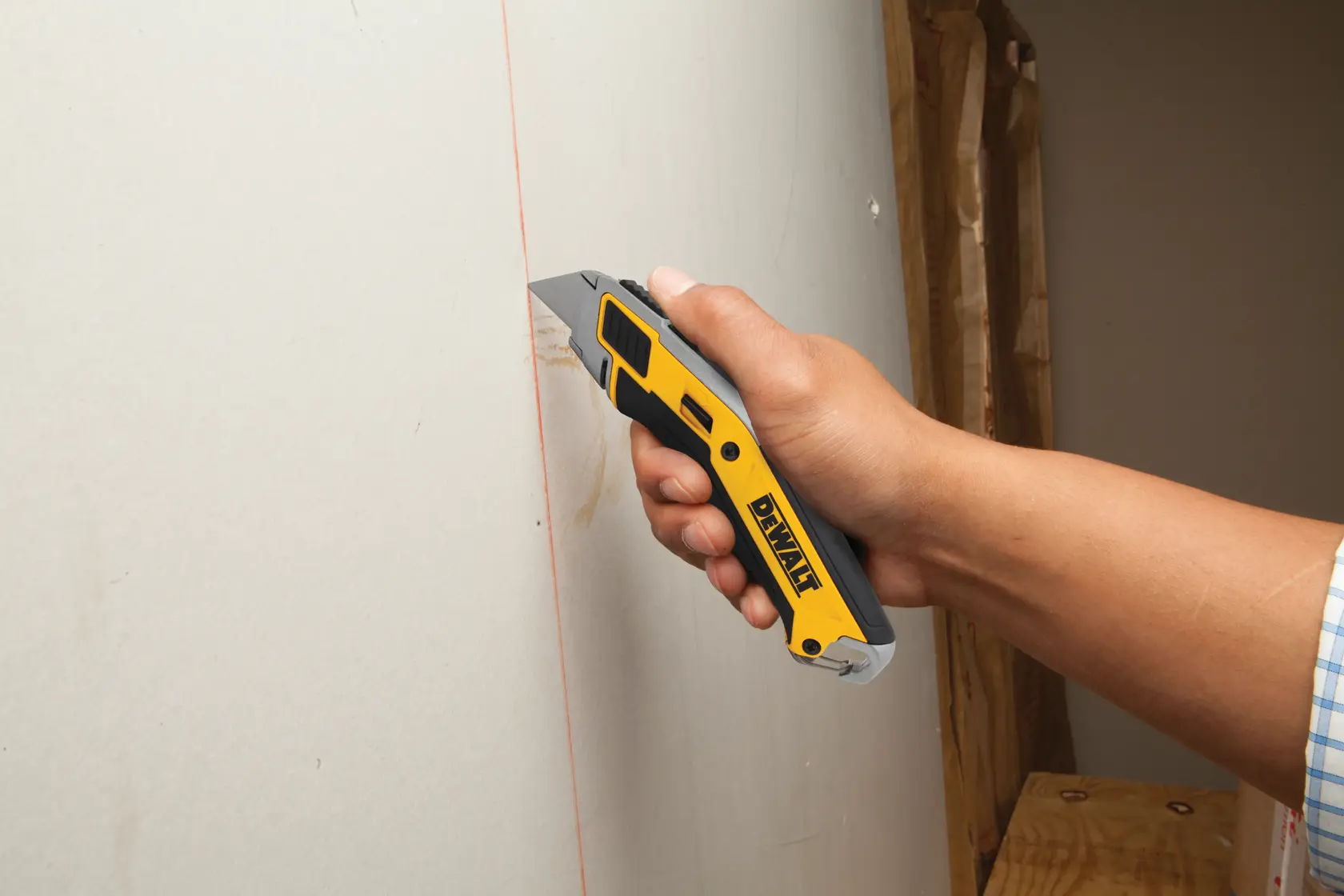 A hand is holding a DEWALT utility knife, cutting along a marked line on a sheet of drywall.