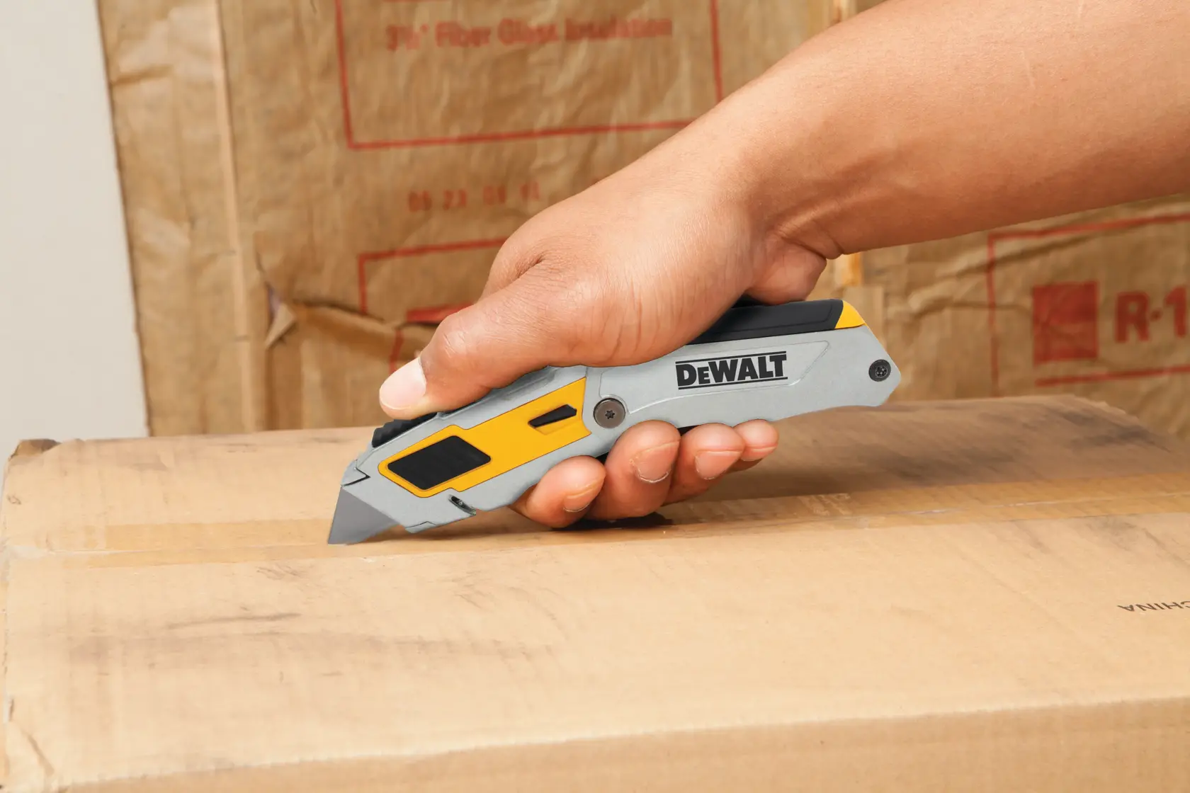 A person using a DEWALT utility knife to cut open a cardboard box. The knife has a gray and yellow handle.