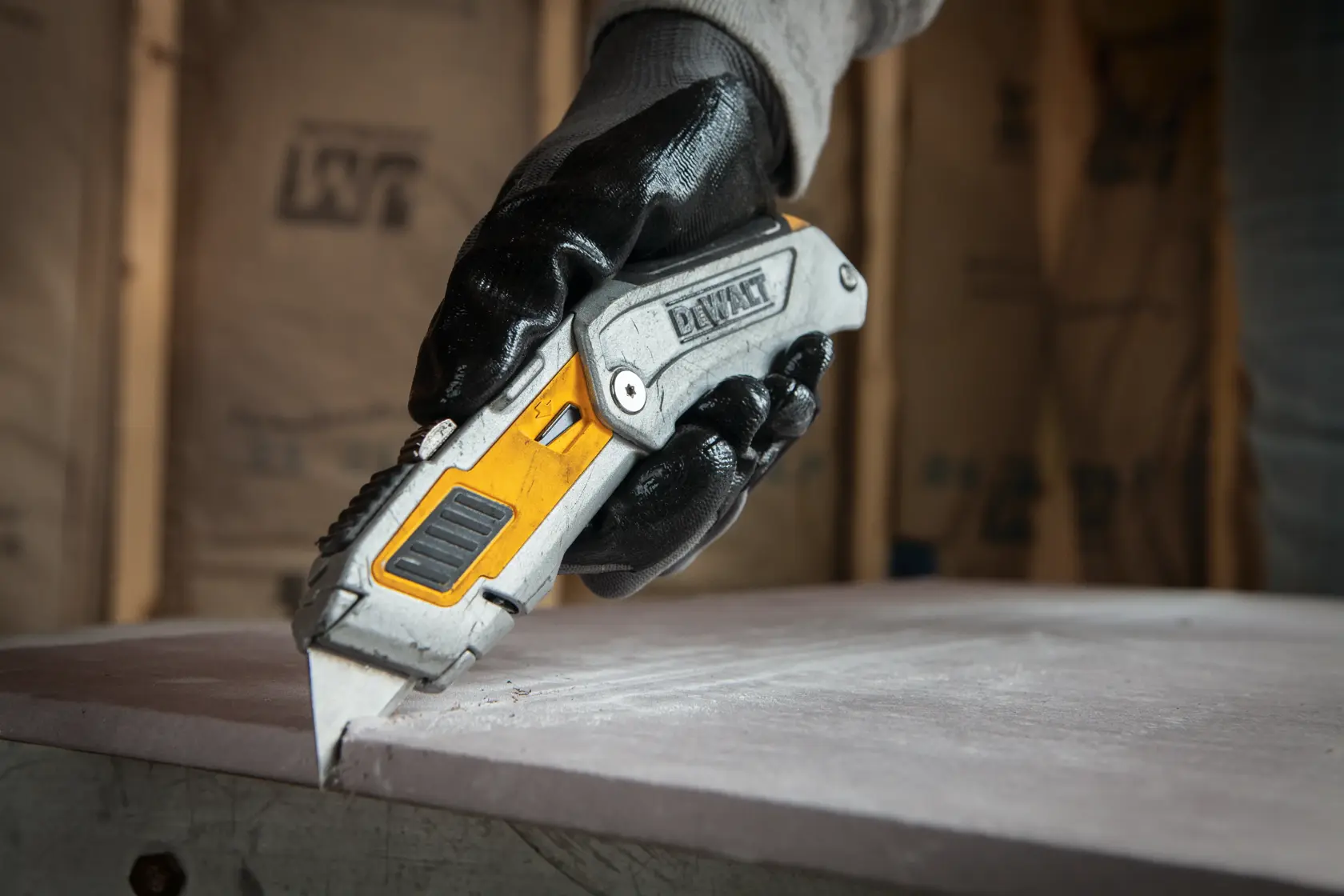 A gloved hand using a DEWALT utility knife to cut a piece of material on a work surface.