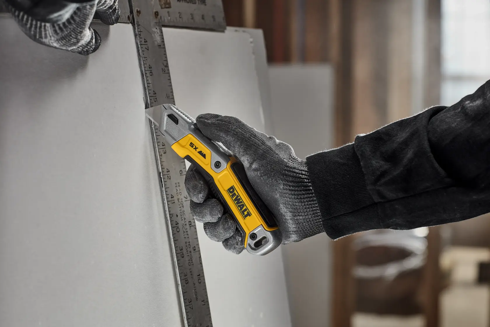 A person wearing protective gloves is using a yellow and silver DEWALT utility knife to cut drywall against a metal ruler.