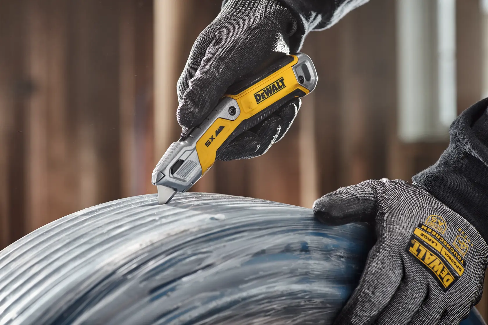A person wearing black and gray protective DEWALT gloves is using a yellow and silver DEWALT utility knife to cut through a wrapped material, possibly plastic or cable, in an industrial setting.