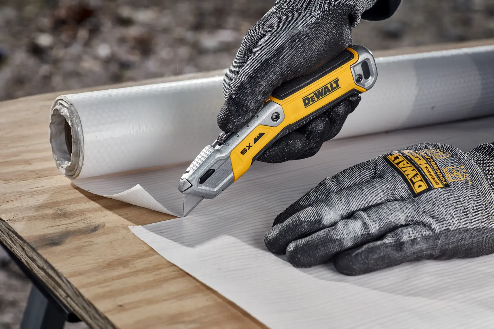 A person wearing protective gloves is using a yellow and silver DEWALT utility knife to cut a sheet of white material on a wooden surface, with a roll of the same material in the background.