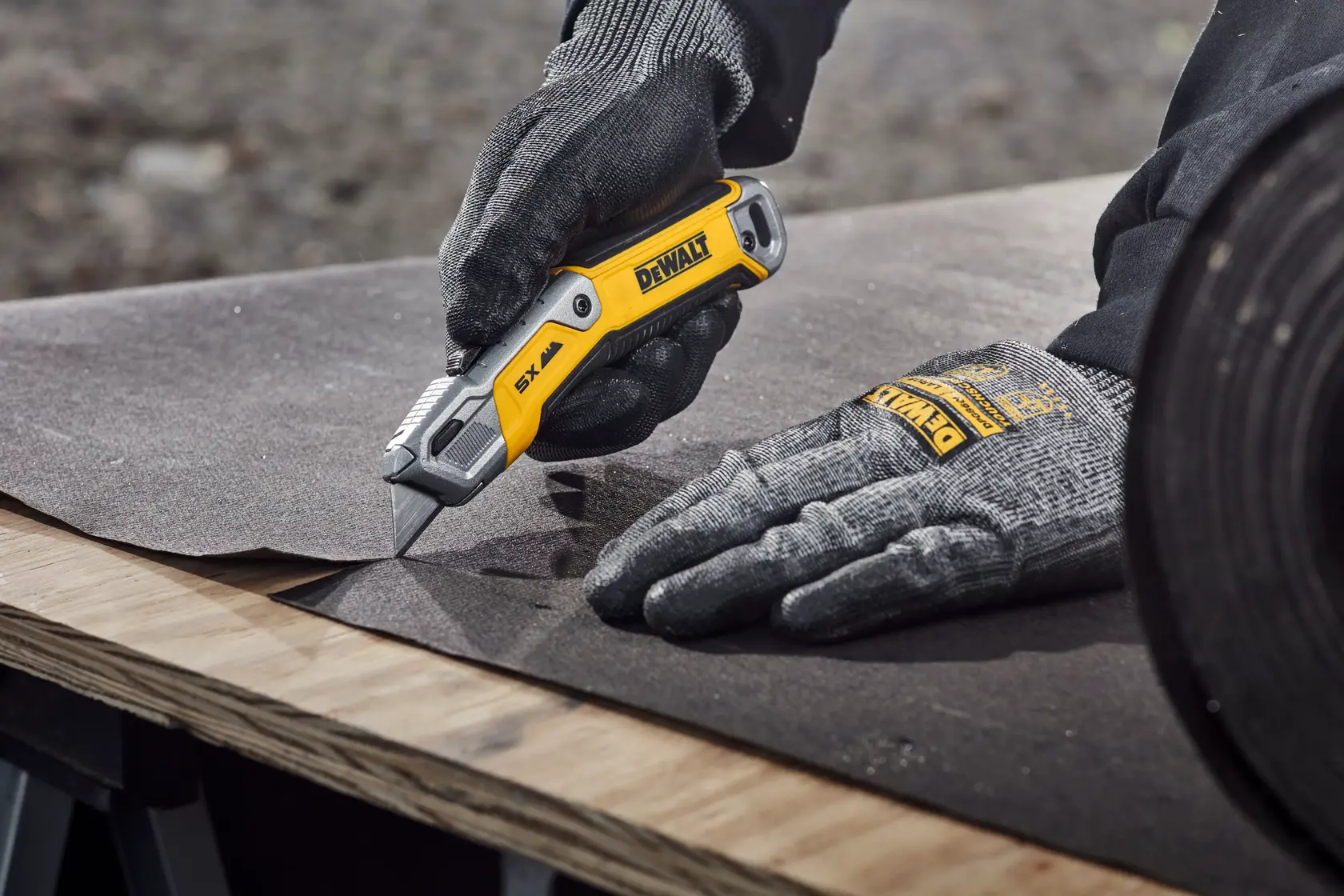 A person wearing gray gloves is using a DEWALT utility knife to cut a sheet of material on a wooden surface.