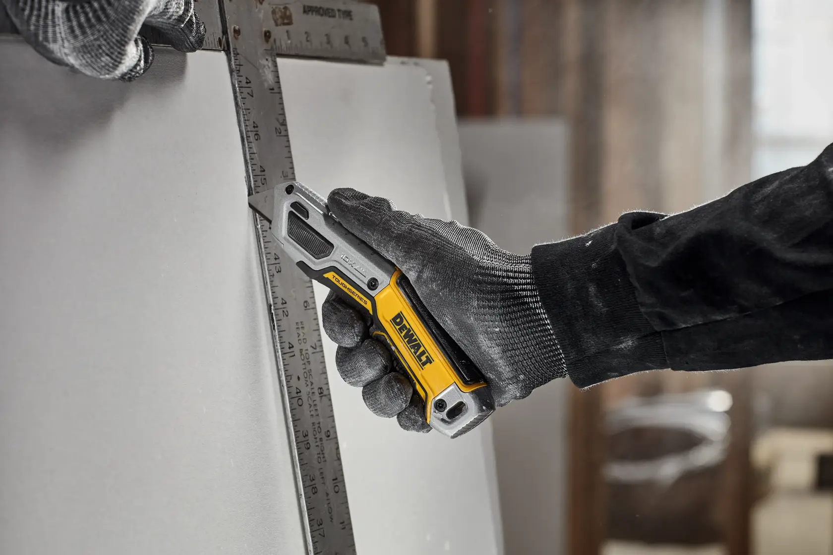 A person wearing work gloves uses a yellow and black DEWALT utility knife to cut a sheet of material along a metal ruler.