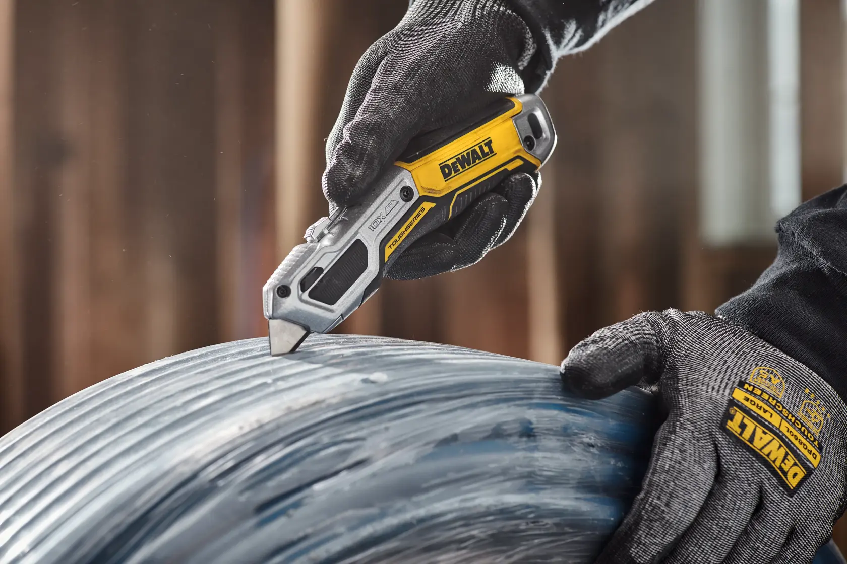 A person wearing DEWALT branded gloves uses a DEWALT folding utility knife to cut through a coil of material in an indoor workshop setting.