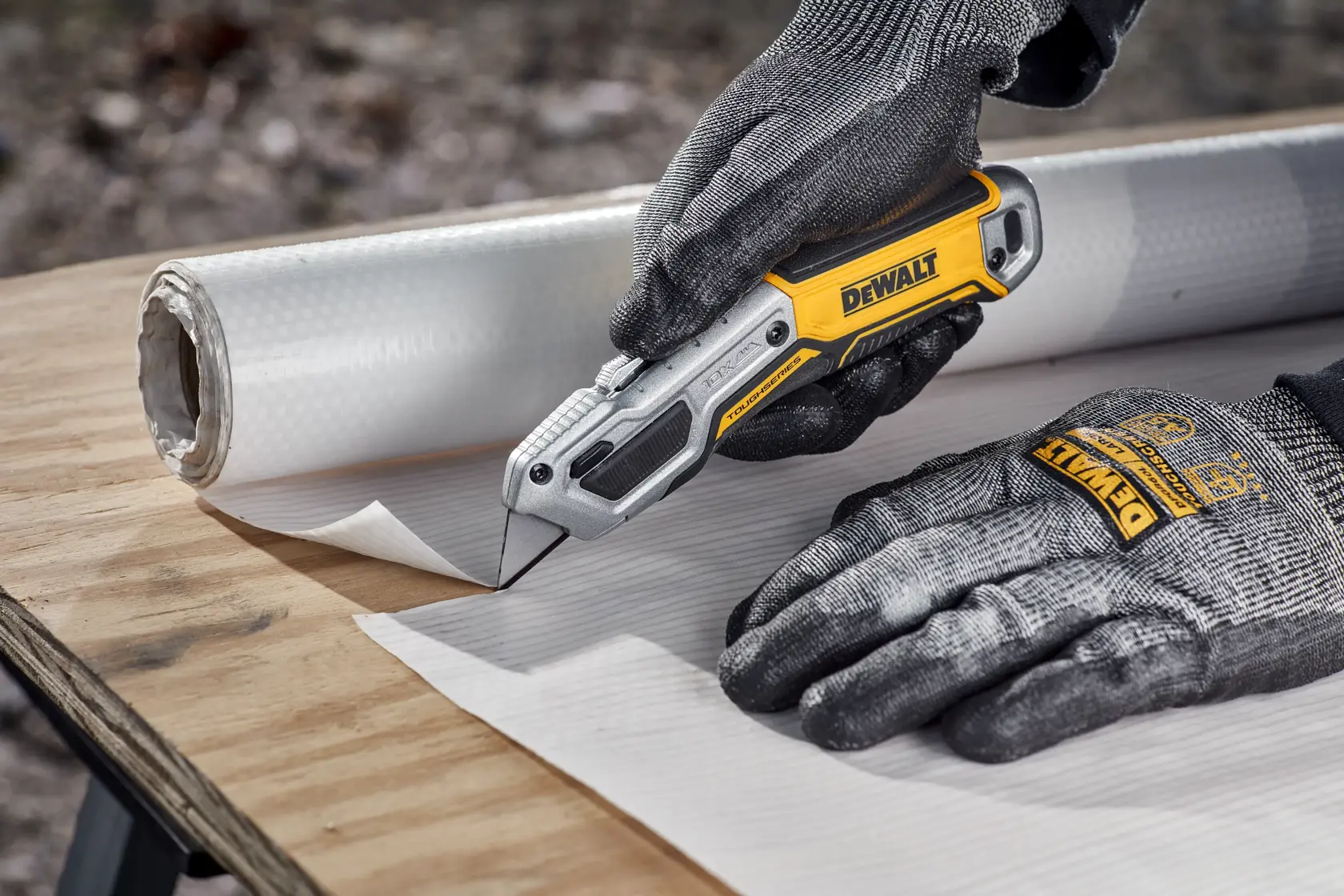 A pair of gloved hands using a DEWALT utility knife to cut a sheet of material on a wooden surface, with a roll of the same material beside.