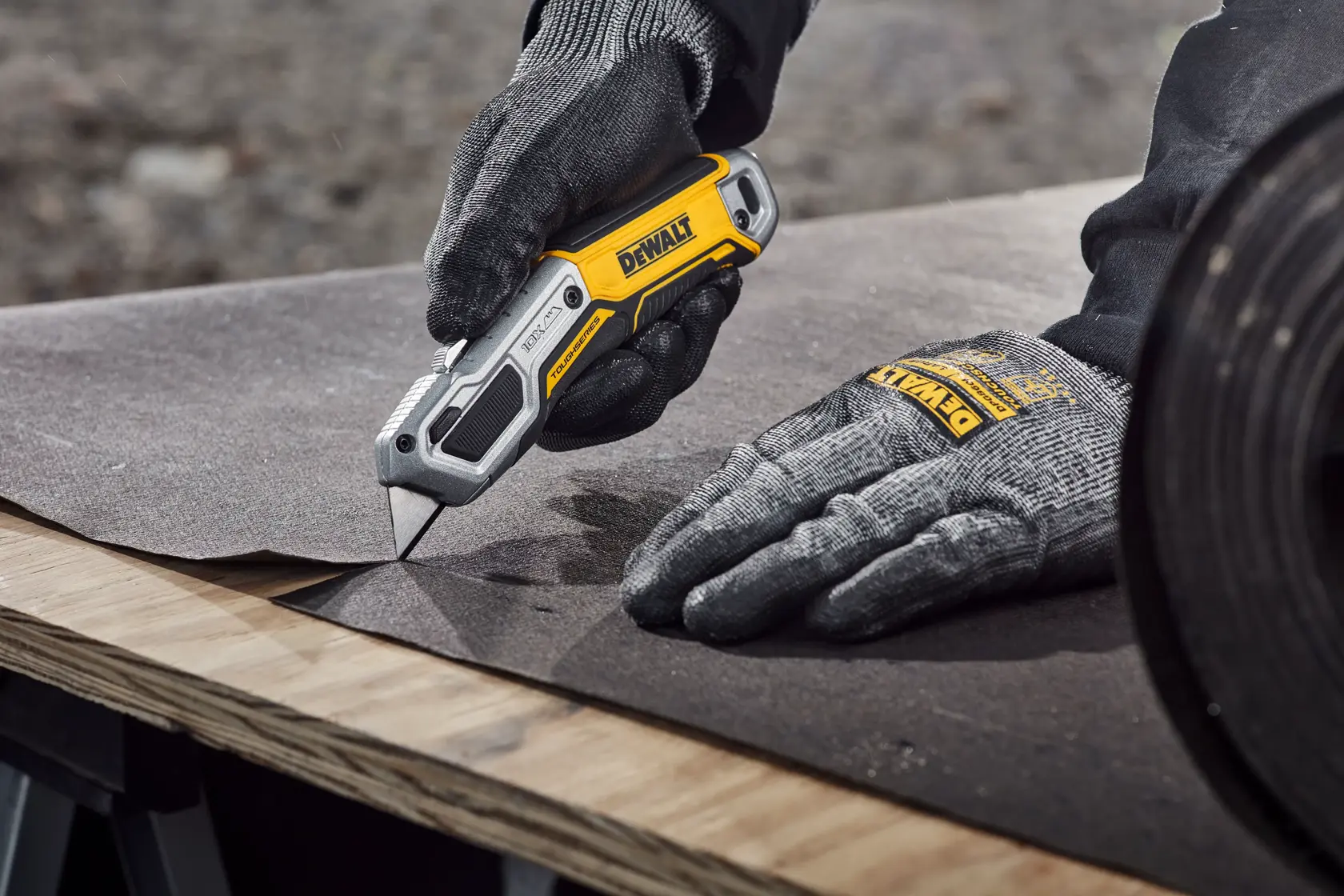 A pair of gloved hands using a DEWALT utility knife to cut a sheet of material on a wooden surface, with a roll of the same material beside.
