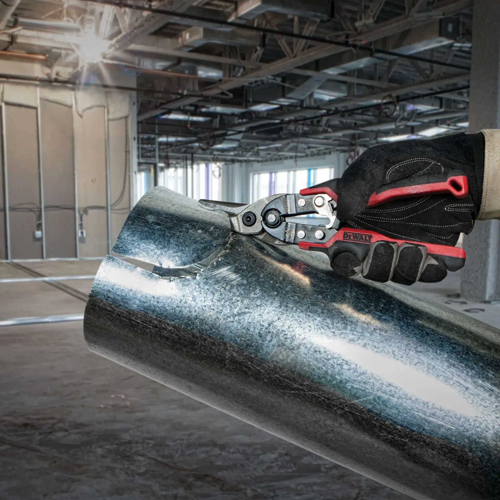 A gloved hand using DEWALT metal snips to cut through a sheet of galvanized metal pipe inside a construction site with exposed ceiling and unfinished walls.