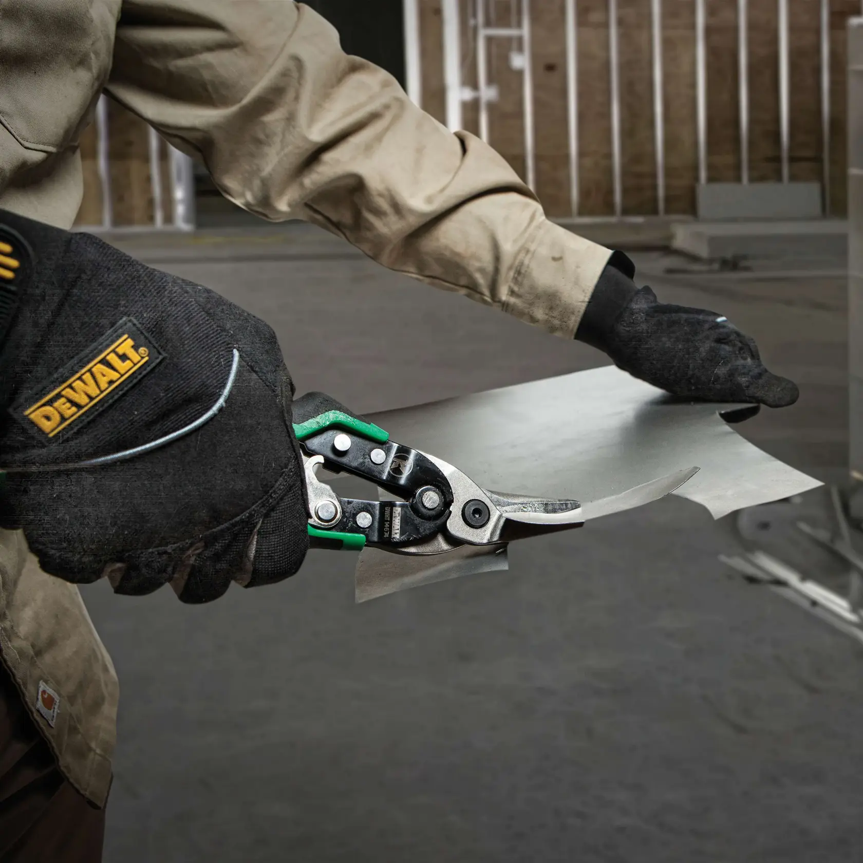 A person wearing black DEWALT gloves uses a pair of DEWALT aviation snips to cut a piece of sheet metal in an indoor workshop setting.