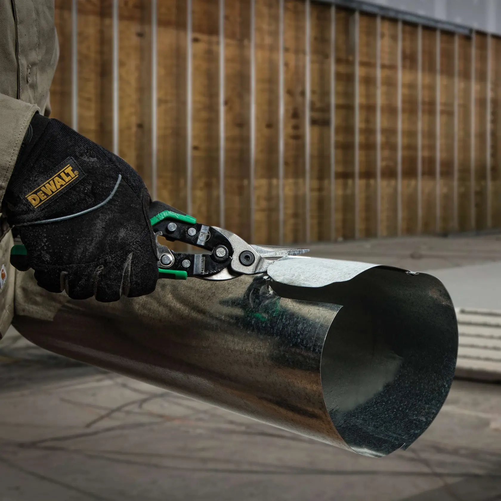 A gloved hand holding DEWALT metal cutting snips, cutting a sheet of metal pipe in an industrial setting.