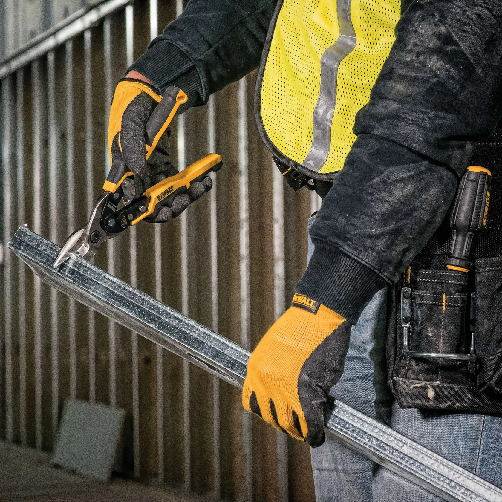 A person wearing work gloves is using a DEWALT metal snip tool to cut a metal stud in a construction setting.
