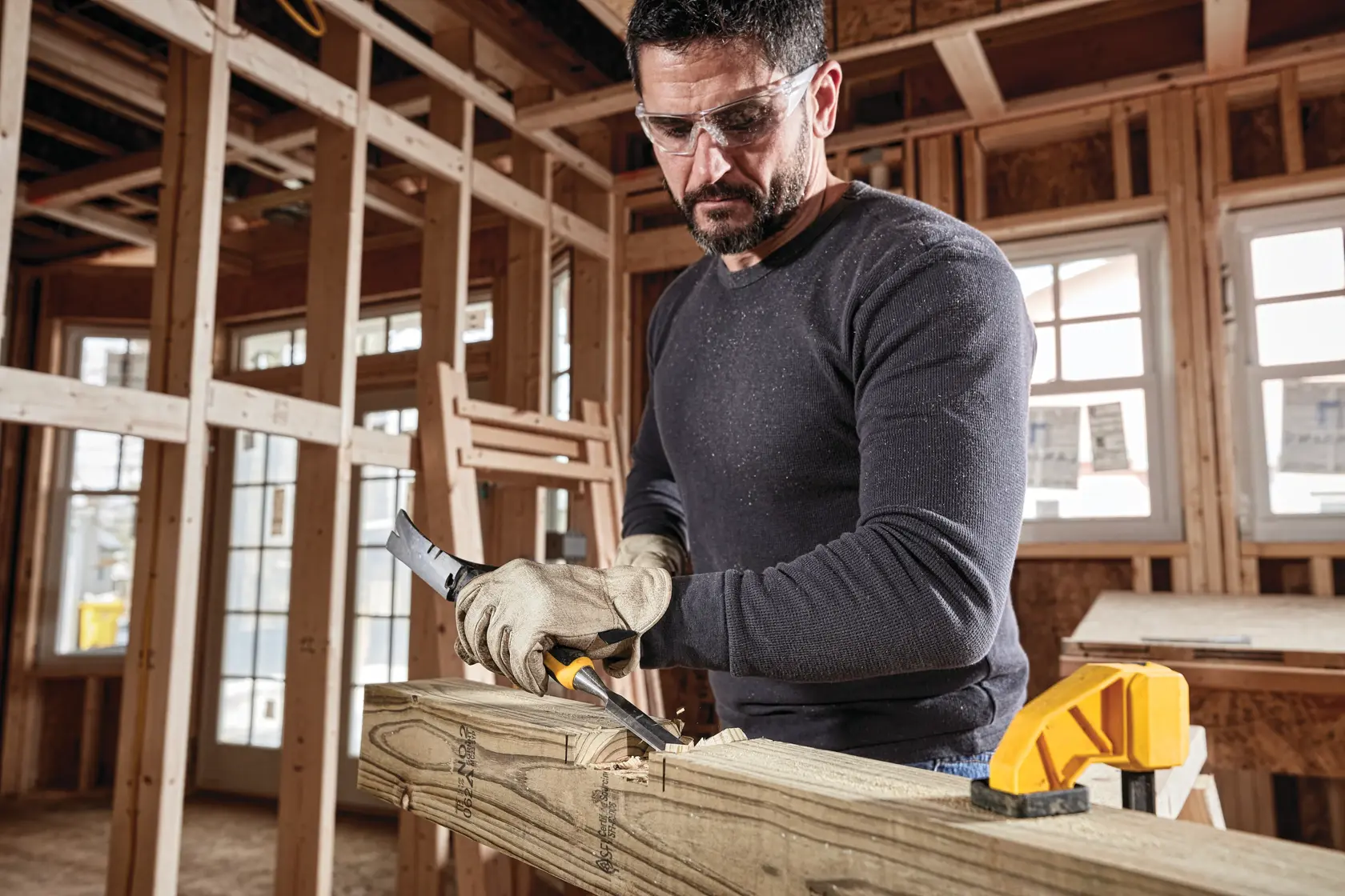 A person wearing gloves is using a DEWALT hand tool to carve wood in a construction site. The scene shows unfinished wooden framing and windows in the background, with a yellow clamp holding the wood in place.