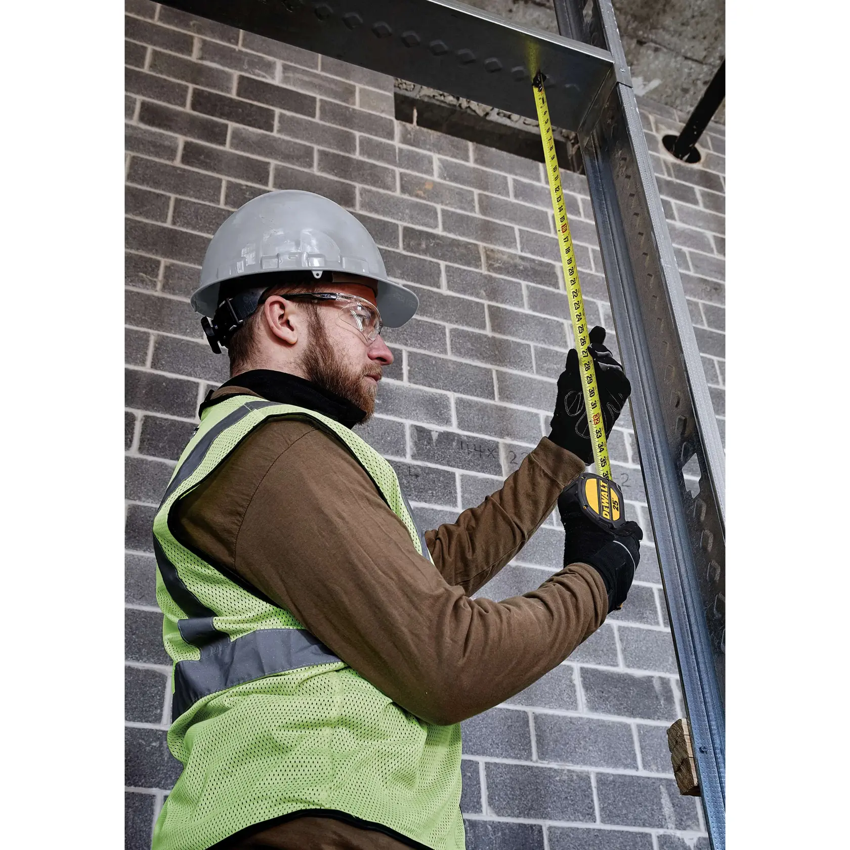 A construction worker wearing a safety vest, gloves, and a hard hat is using a DEWALT tape measure to measure the height of a metal beam against a brick wall inside a building under construction.