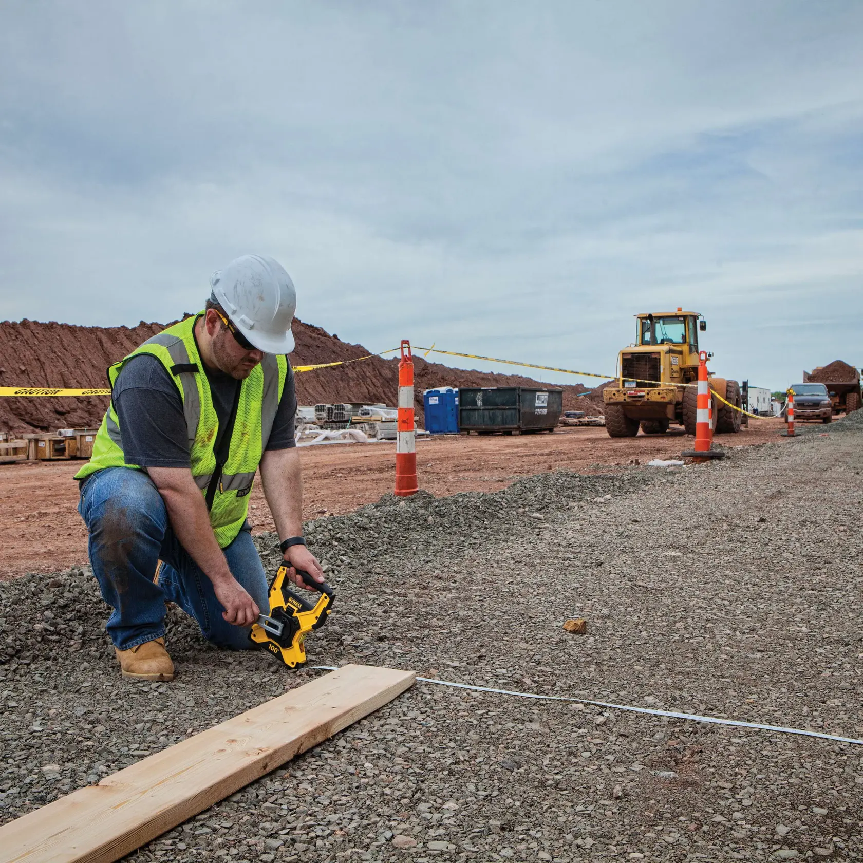 A person wearing a safety vest and hard hat is using a DEWALT measuring tape reel (SKU: DWHT34145) to measure distance at a construction site, with construction vehicles and equipment visible in the background.