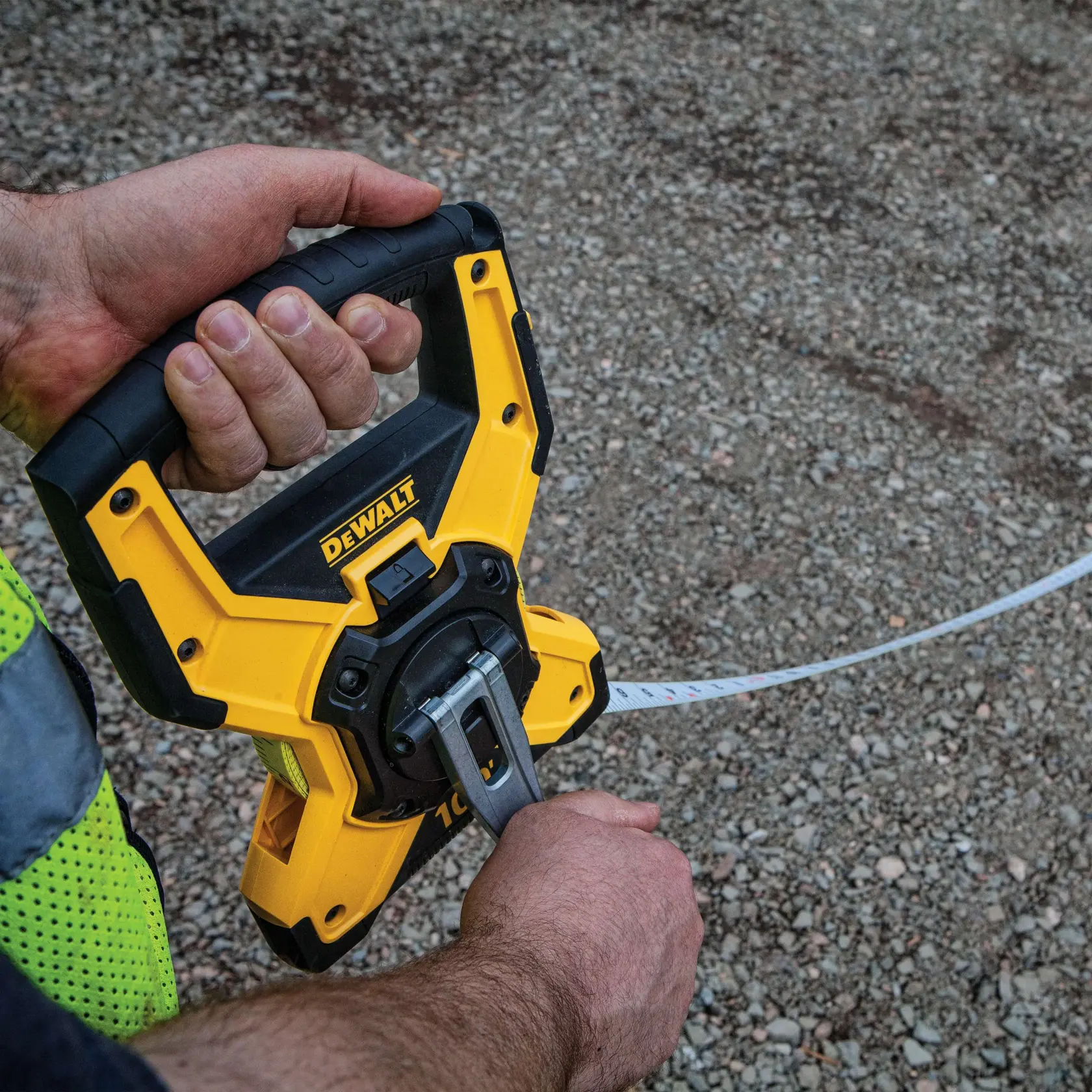 Close-up of a person holding and operating a DEWALT measuring tape reel, using the handle and crank to extend the tape over a gravel surface.