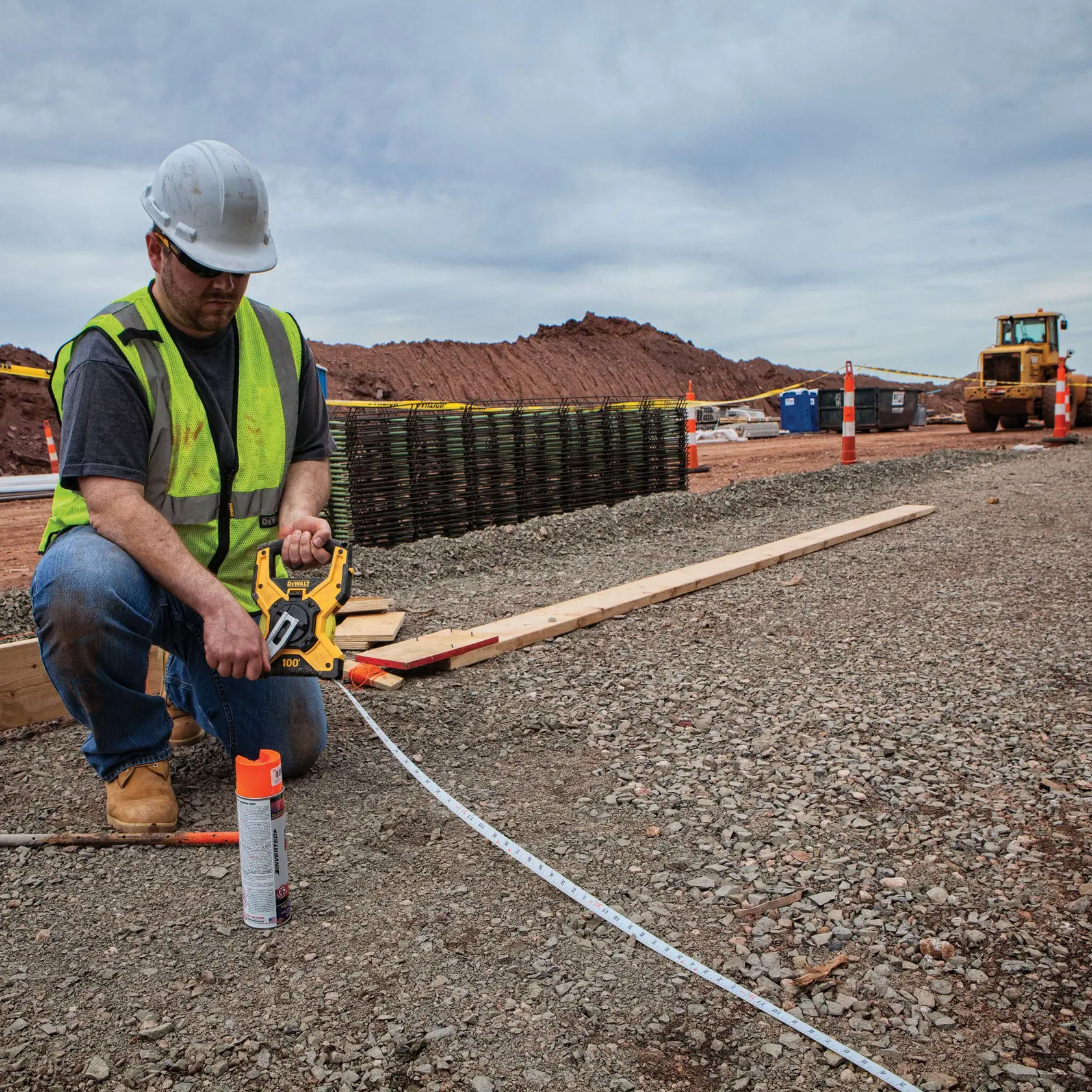 A construction worker kneels outdoors on a gravel surface, wearing a safety vest and hard hat. The worker is using a DEWALT measuring tape reel (SKU: DWHT34145) to measure a distance, with the tape extended along the ground. Construction materials, equipment, and traffic cones are visible in the background.