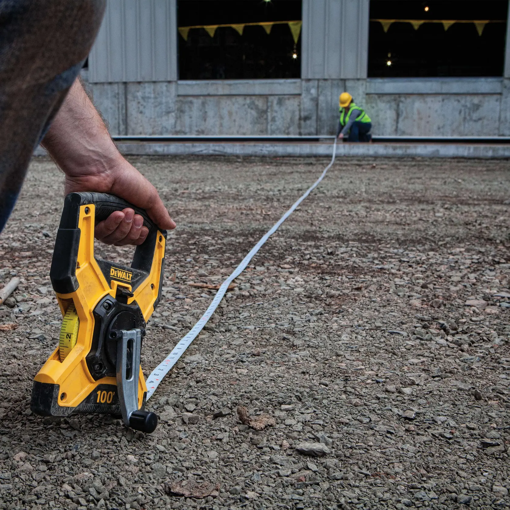 A person uses a DEWALT long measuring tape to measure distance on a rough construction site. Another worker in a yellow hard hat and safety vest is seen at the far end of the tape.
