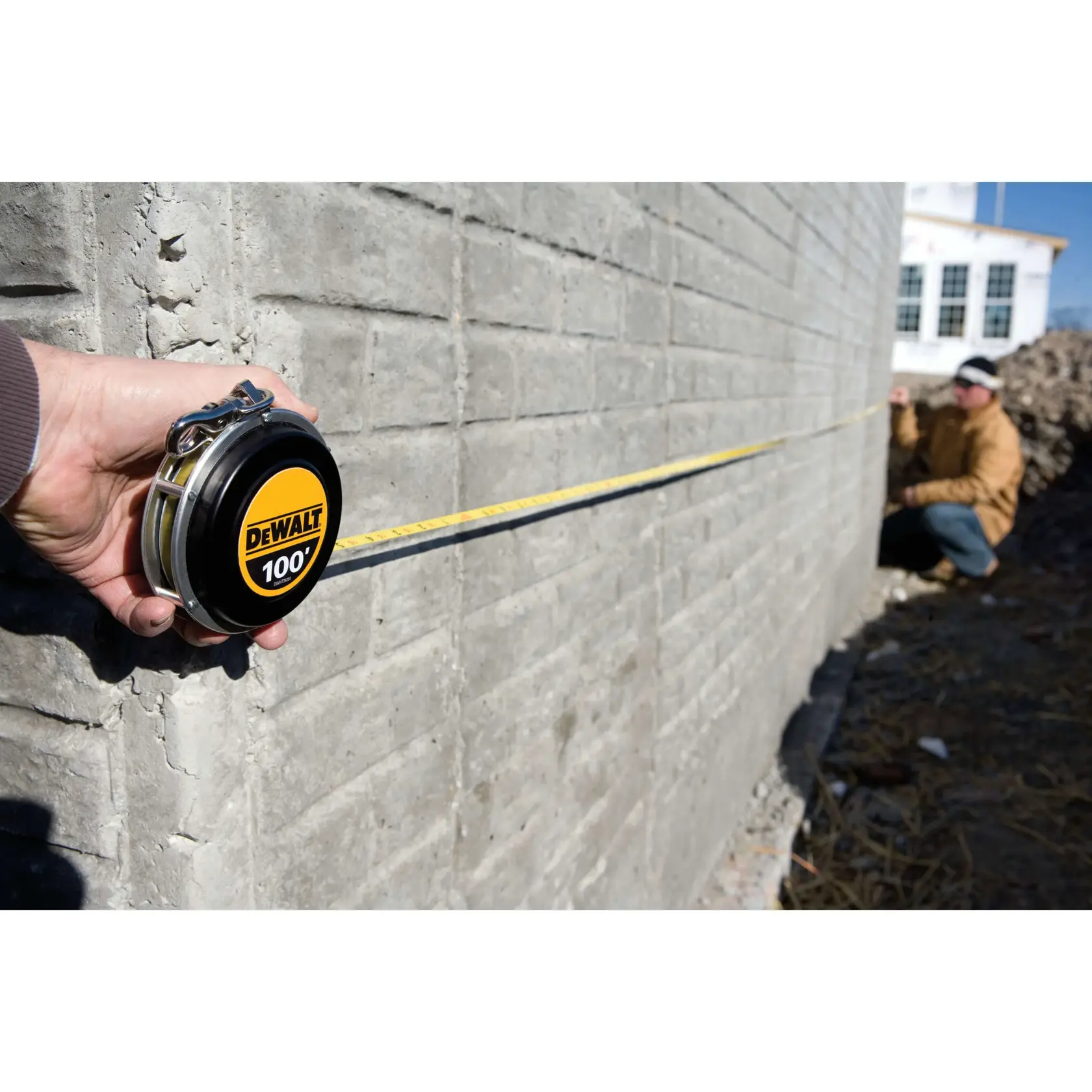 A person uses a DEWALT 100-foot measuring tape to measure the length of a concrete wall at a construction site. Another person is seen in the background holding the tape near the wall.