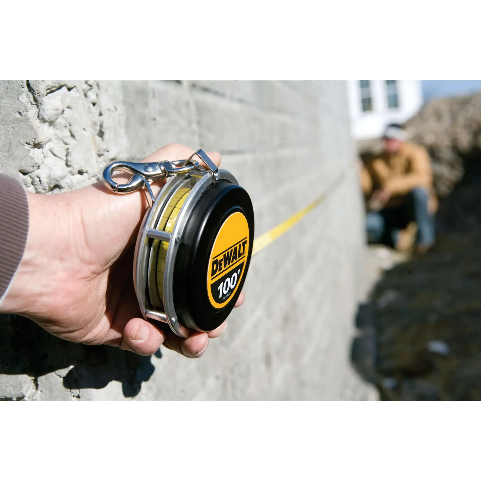 A close-up of a hand holding a DEWALT 100-foot tape measure against a concrete wall, with a yellow measuring tape extended towards another person in the background.