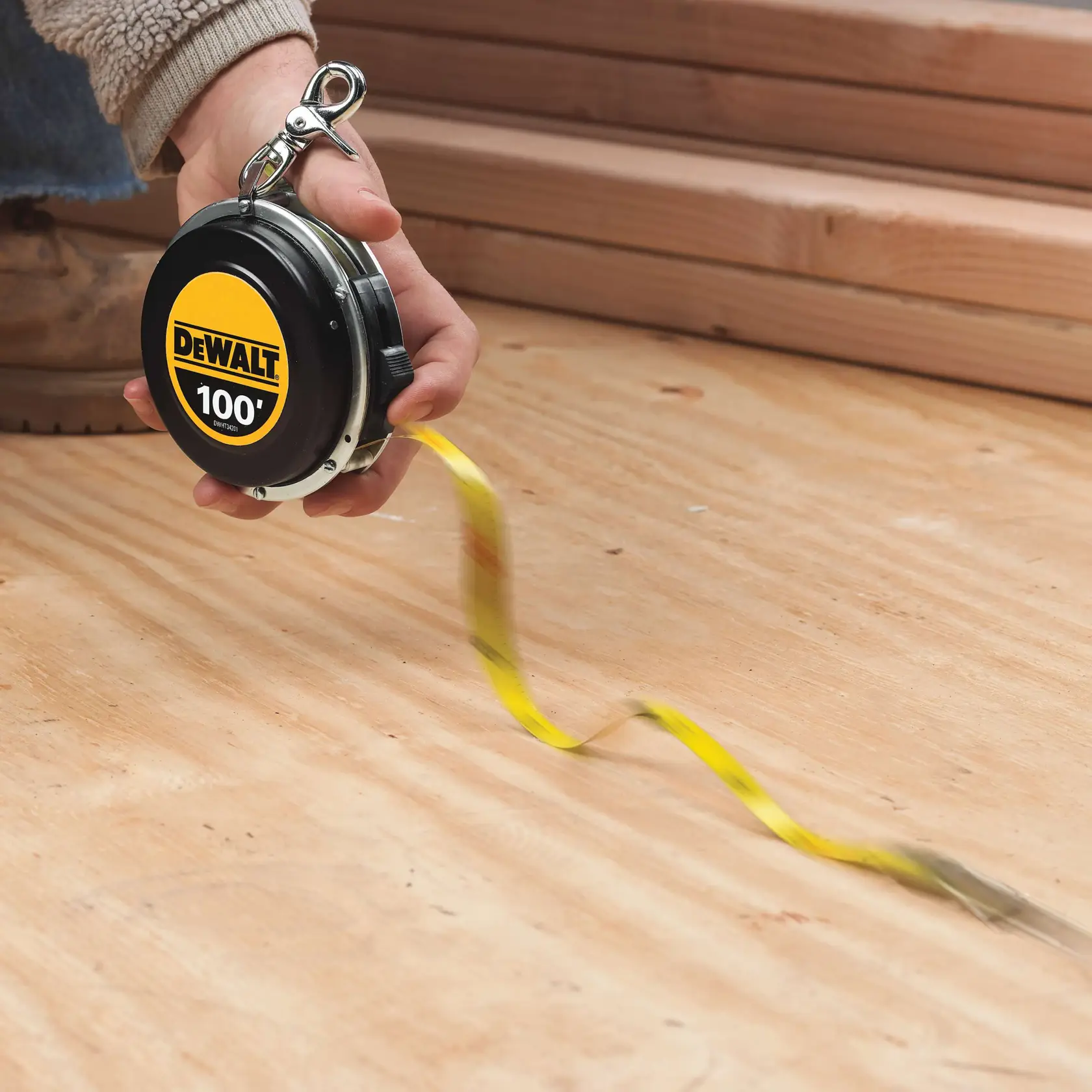 A person is holding a DEWALT 100-foot tape measure, with the tape extended on a wooden floor. There are stacked wooden planks in the background.