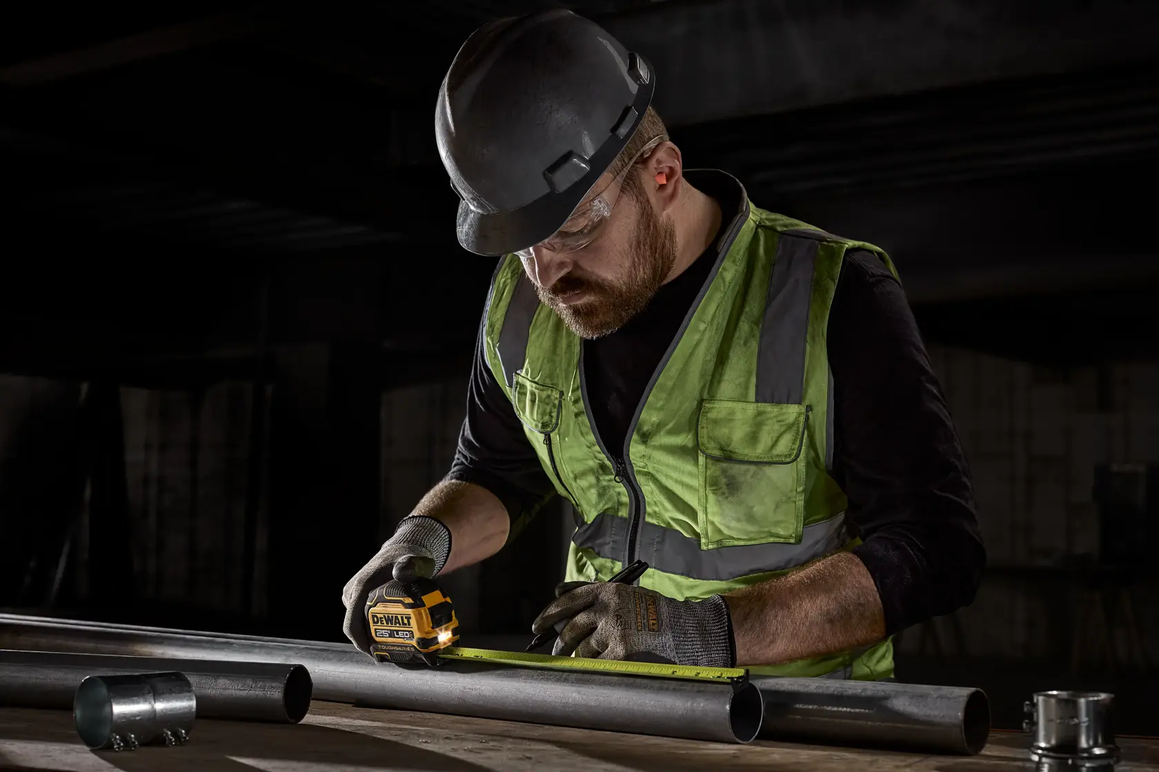 A construction worker wearing a hard hat, safety vest, and gloves is using a DEWALT tape measure to measure a metal pipe in an industrial setting.