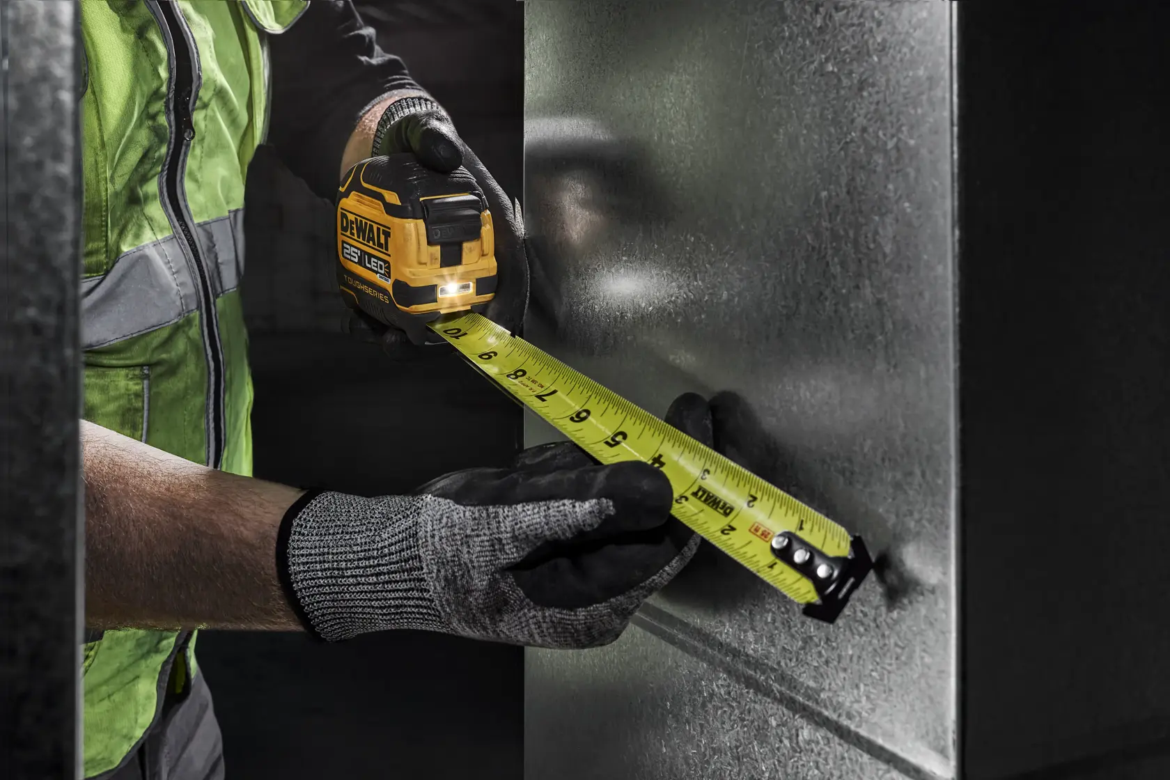 A person wearing safety gloves and a high-visibility vest uses a yellow DEWALT tape measure with LED light to measure the length of a metal surface.