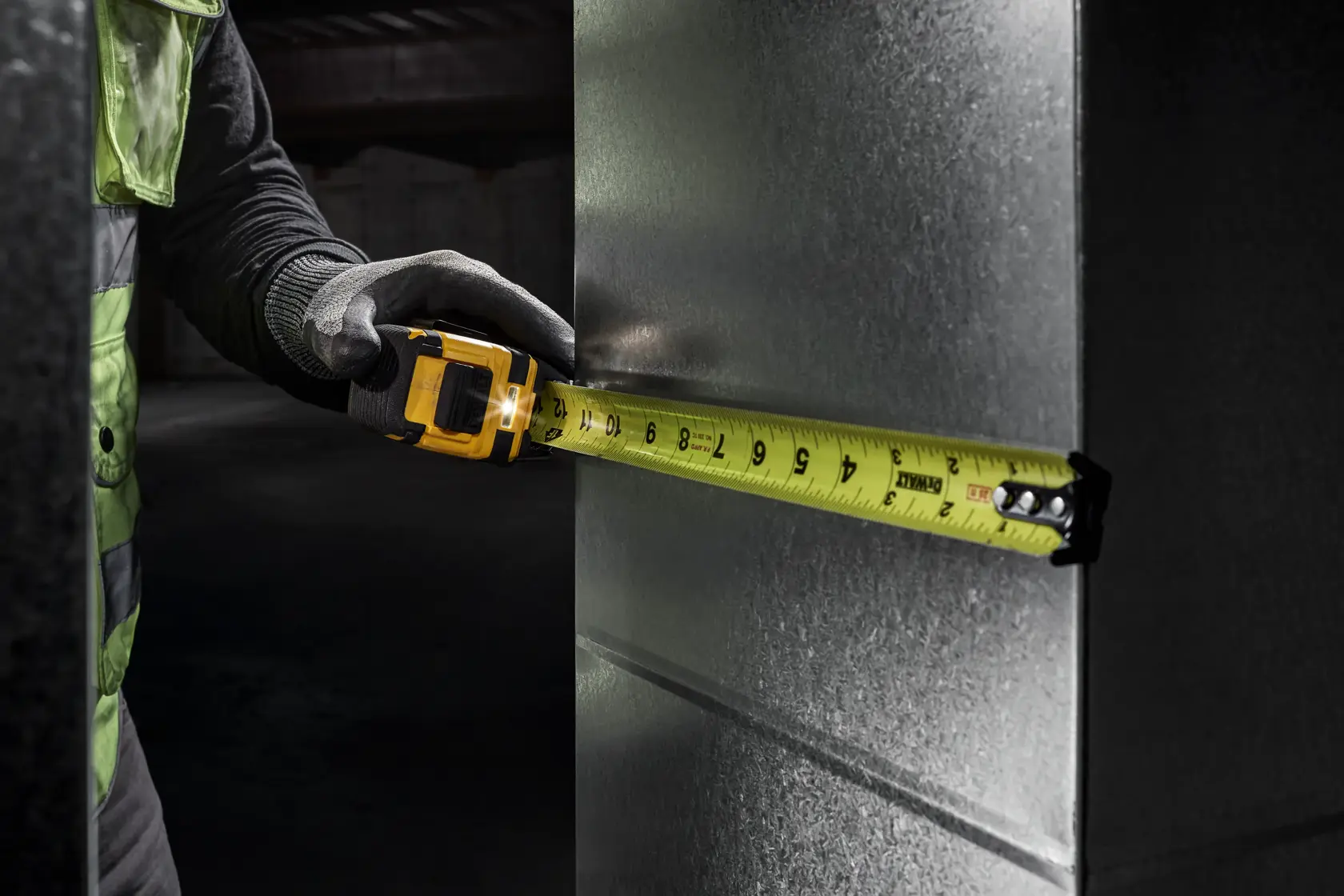 A person wearing gloves uses a yellow DEWALT tape measure with an integrated LED light to measure a metal surface. The image highlights improved visibility due to the LED light, with text at the bottom reading 'Improved Visibility with Powerful Integrated LED Light.'