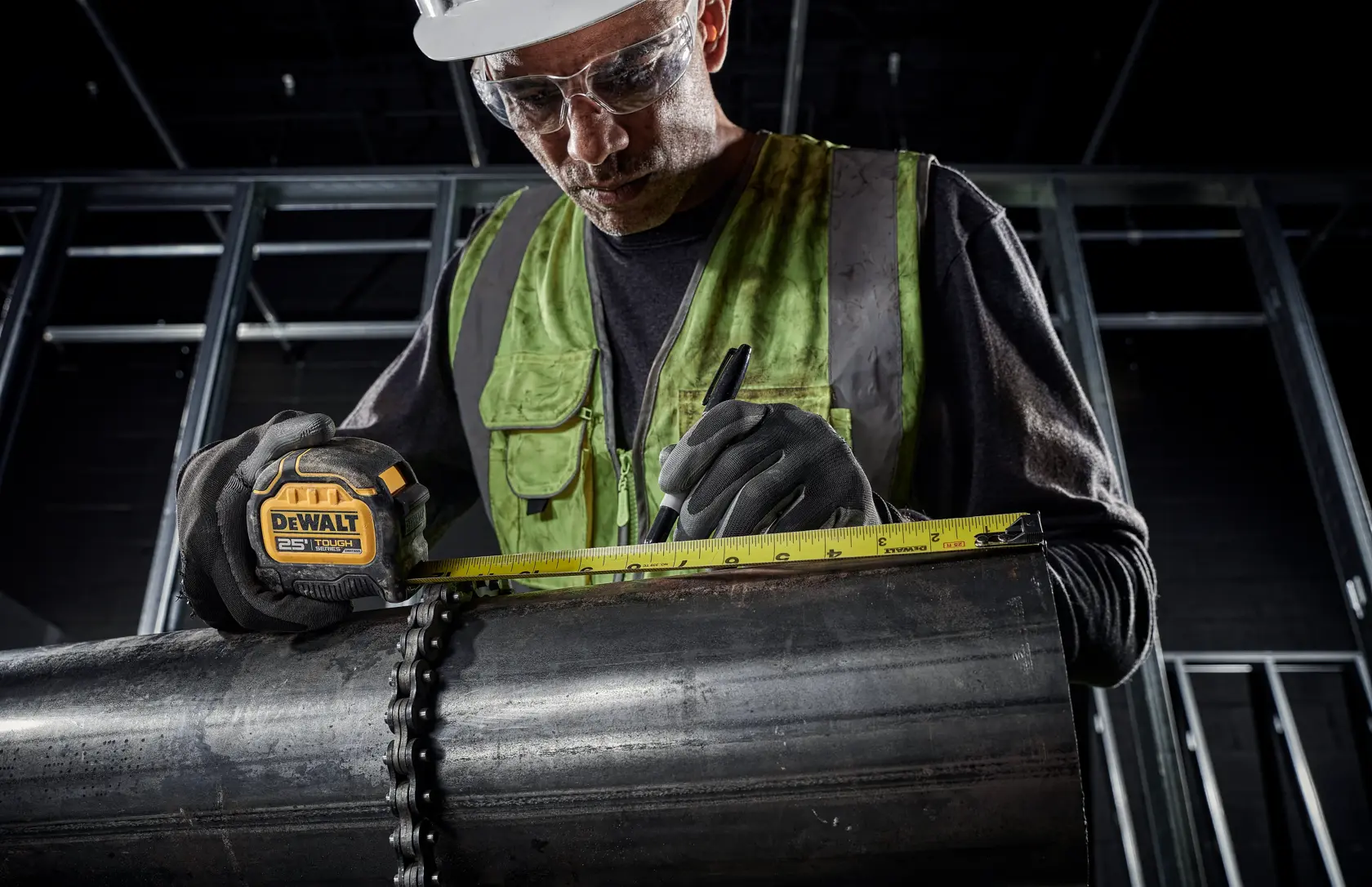A person wearing a green safety vest and gloves uses a DEWALT 25 ft tape measure to measure a large metal pipe in an industrial setting. The individual marks measurements on the pipe with a black marker.