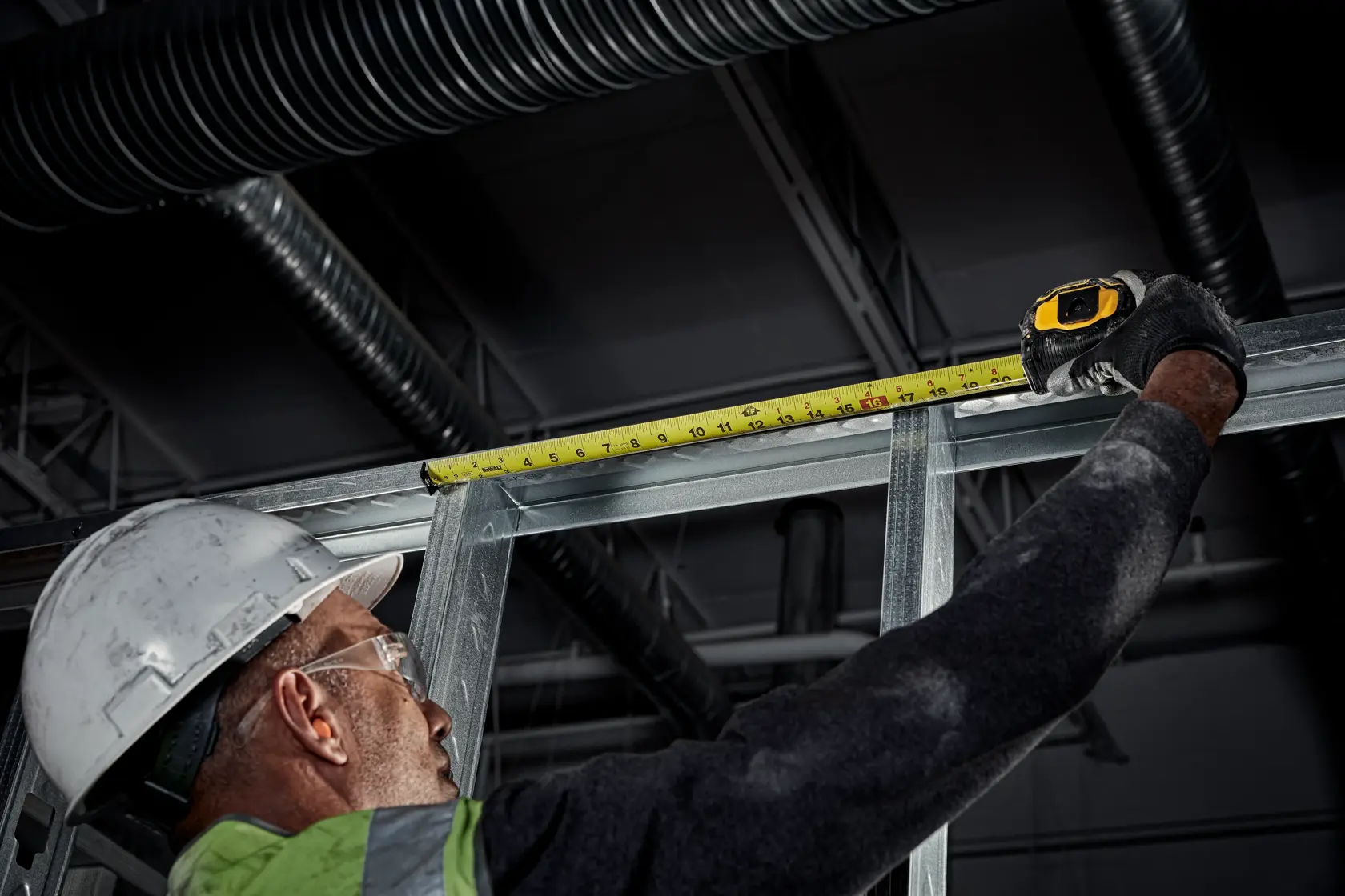 A person wearing a hard hat and safety vest uses a DEWALT tape measure to measure the length of a metal frame in an industrial setting.